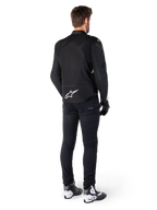 Alpinestars T-Jaws V4 Wasserdichte Jacke, Schwarz und Weiß, Rückansicht mit weißem Stern-Logo auf dem unteren Rücken, getragen von einem Model mit schwarzer Motorradhose, schwarzen Handschuhen und weiß-schwarzen Motorradstiefeln