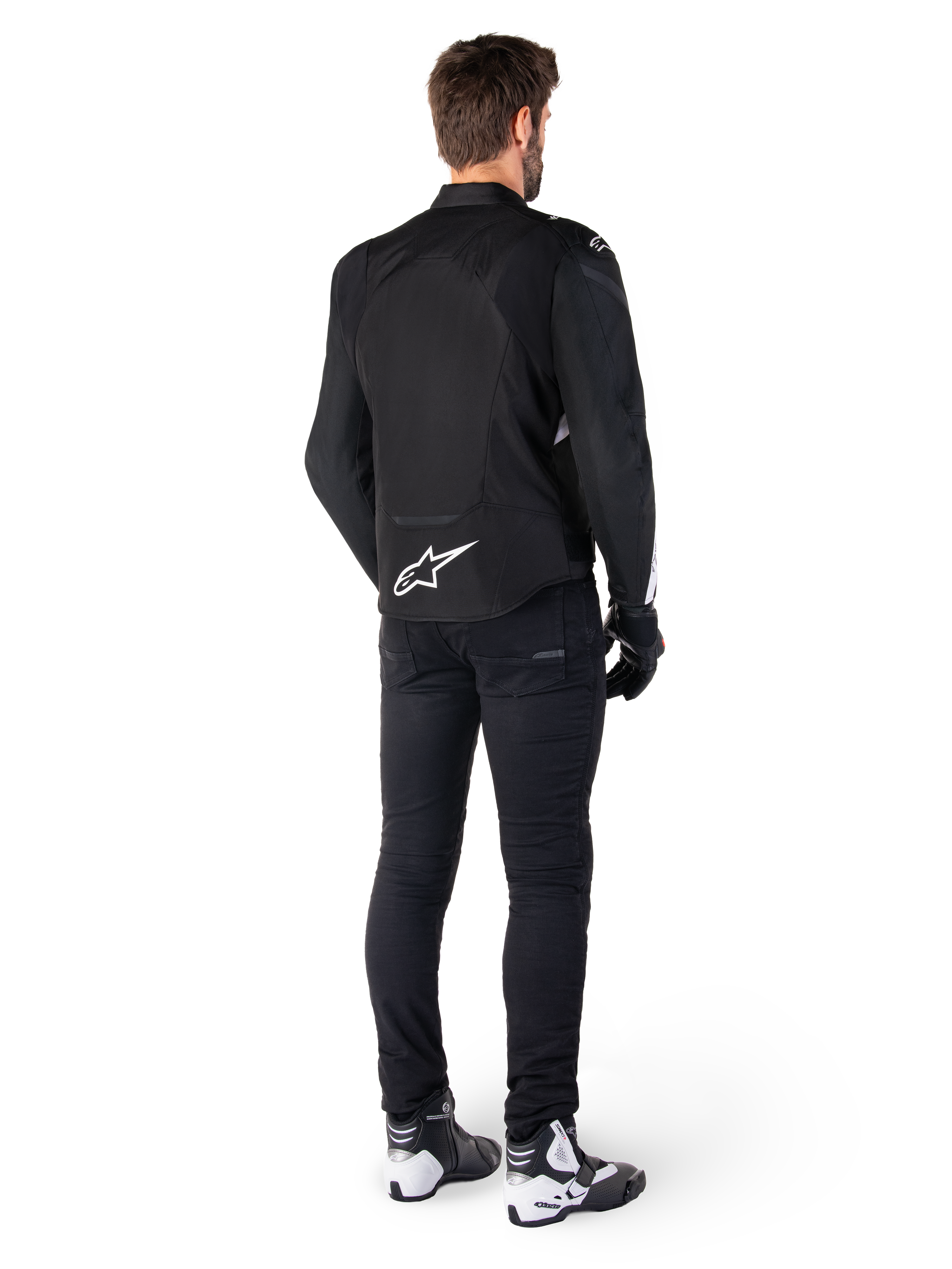 Alpinestars T-Jaws V4 Wasserdichte Jacke, Schwarz und Weiß, Rückansicht mit weißem Stern-Logo auf dem unteren Rücken, getragen von einem Model mit schwarzer Motorradhose, schwarzen Handschuhen und weiß-schwarzen Motorradstiefeln