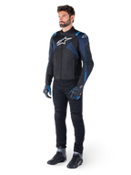 Alpinestars T-Jaws V4 Wasserdichte Jacke in Schwarz, Dunkelblau und Blithe-Blau, getragen von einem Model mit schwarzer Motorradhose und Motorradstiefeln, mit weißem Alpinestars-Logo auf der Brust und blauen Schulterakzenten, sportliche Fahrausrüstung.