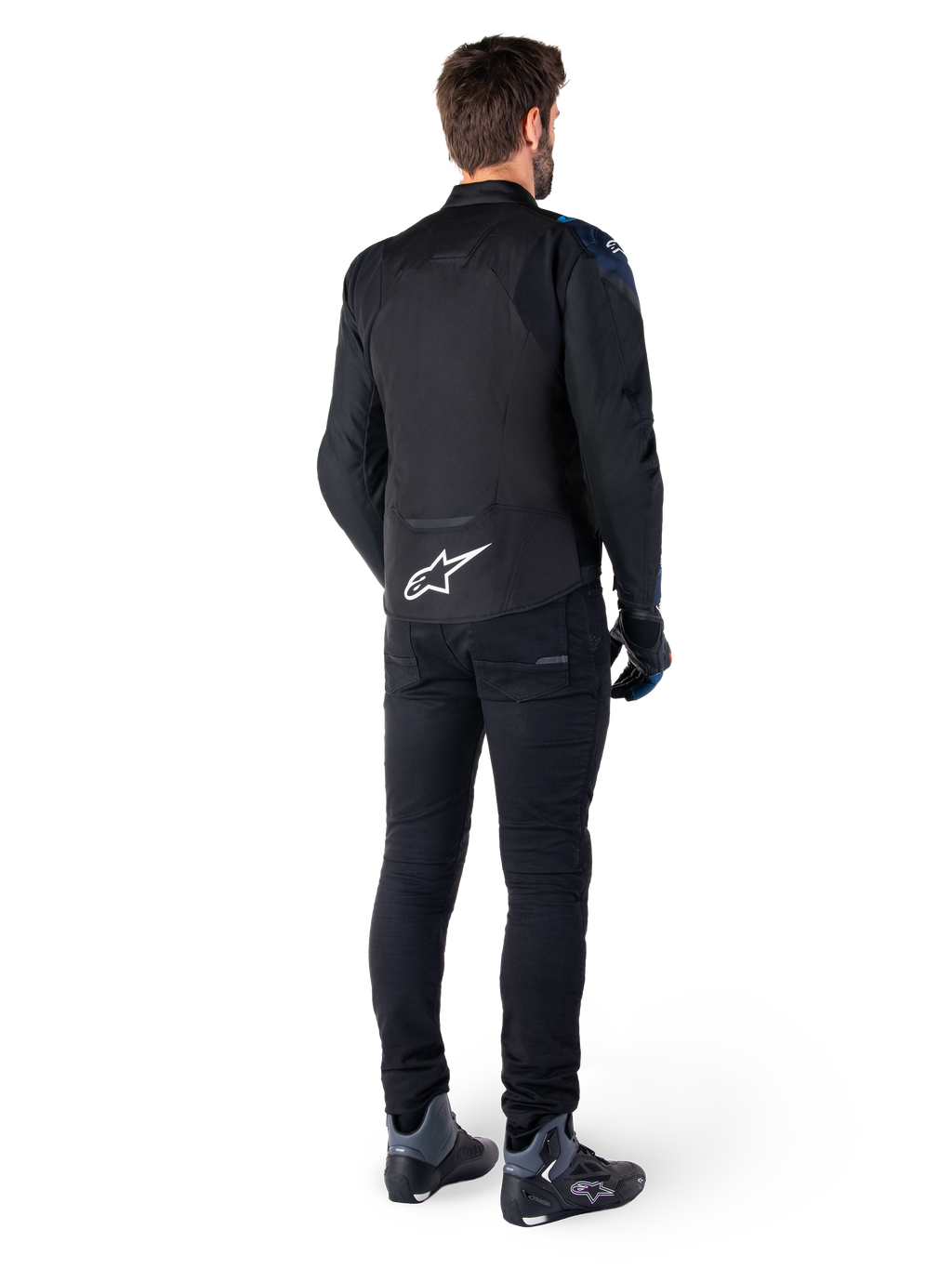 Rückansicht eines Mannes, der die Alpinestars T-Jaws V4 Wasserdichte Jacke in Schwarz, Dunkelblau und Blithe Blue trägt, mit einem markanten weißen Astar-Logo auf dem unteren Rücken, kombiniert mit einer schwarzen Hose und Motorradschuhen.