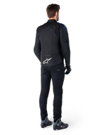 Rückansicht eines Mannes, der die Alpinestars T-Jaws V4 Wasserdichte Jacke in Schwarz, Dunkelblau und Blithe Blue trägt, mit einem markanten weißen Astar-Logo auf dem unteren Rücken, kombiniert mit einer schwarzen Hose und Motorradschuhen.