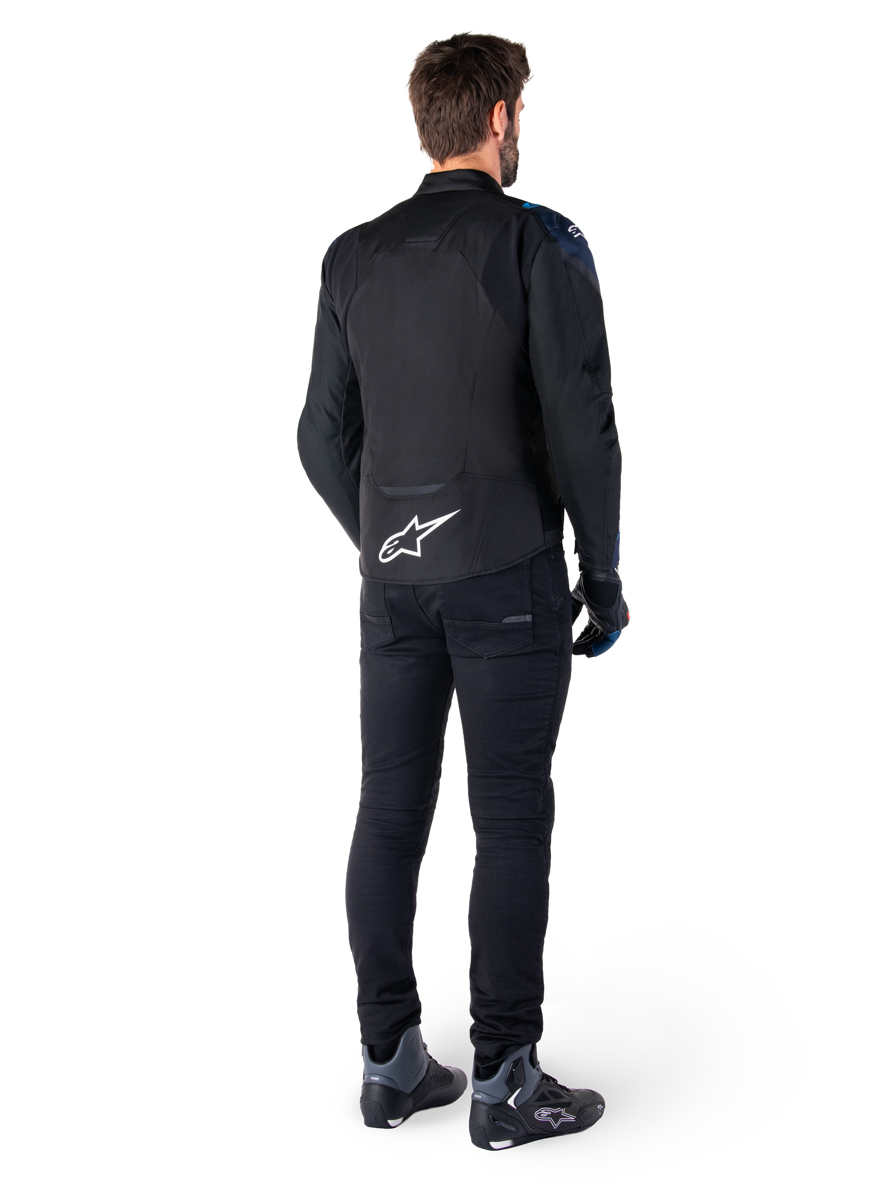 Rückansicht eines Mannes, der die Alpinestars T-Jaws V4 Wasserdichte Jacke in Schwarz, Dunkelblau und Blithe Blue trägt, mit einem markanten weißen Astar-Logo auf dem unteren Rücken, kombiniert mit einer schwarzen Hose und Motorradschuhen.