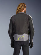 Alpinestars T-Jaws V4 Wasserdichte Jacke, Schwarz Eisgrau Dunkelgrau, Rückansicht am Model, mit einem hellgrauen Einsatz am unteren Rücken und einem markanten neongelben Stern-Logo, mehrlagige Textilkonstruktion, sportliche Silhouette, getragen mit schwarzen Handschuhen und einer dunklen Hose.