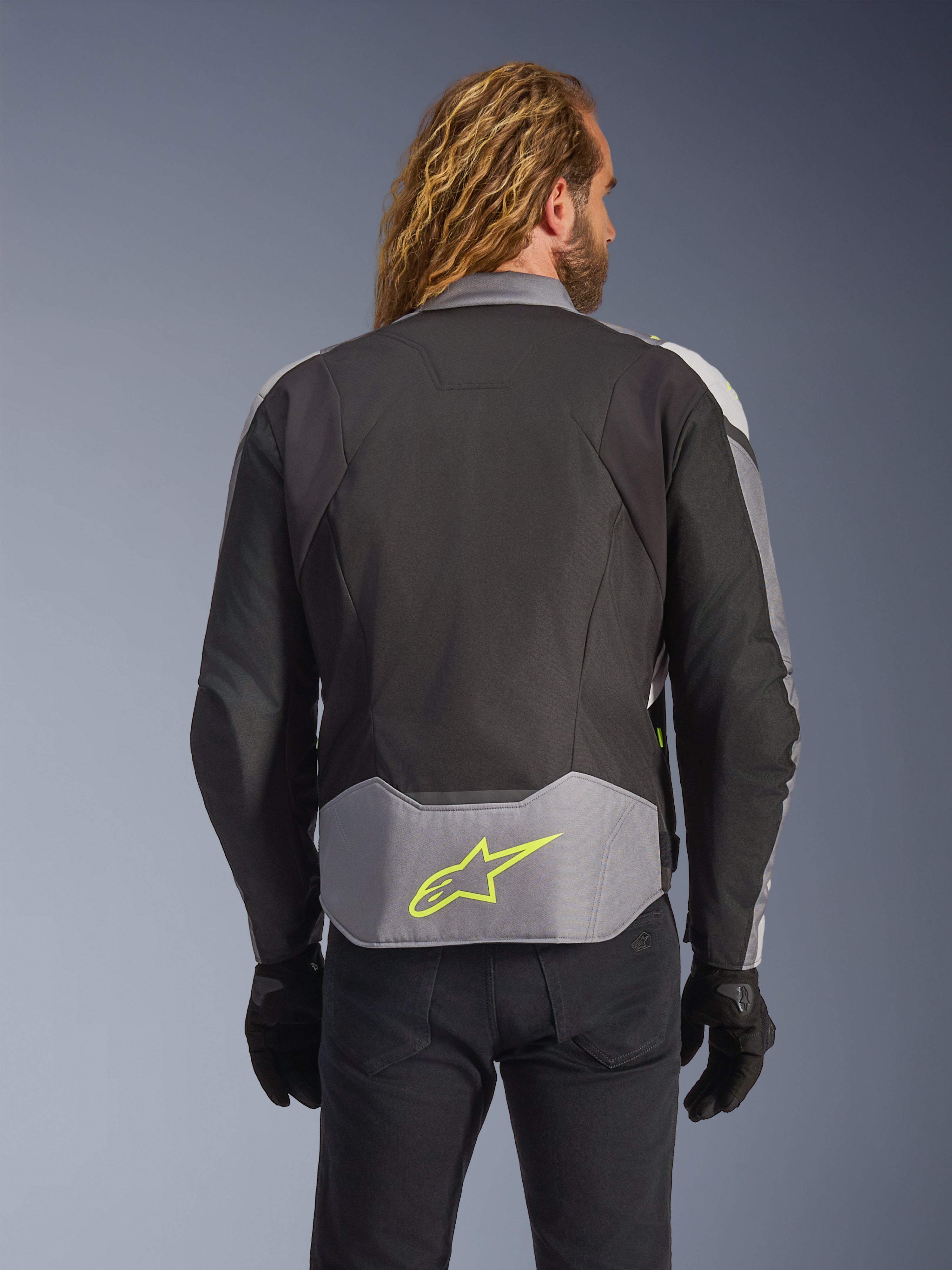 Alpinestars T-Jaws V4 Wasserdichte Jacke, Schwarz Eisgrau Dunkelgrau, Rückansicht am Model, mit einem hellgrauen Einsatz am unteren Rücken und einem markanten neongelben Stern-Logo, mehrlagige Textilkonstruktion, sportliche Silhouette, getragen mit schwarzen Handschuhen und einer dunklen Hose.