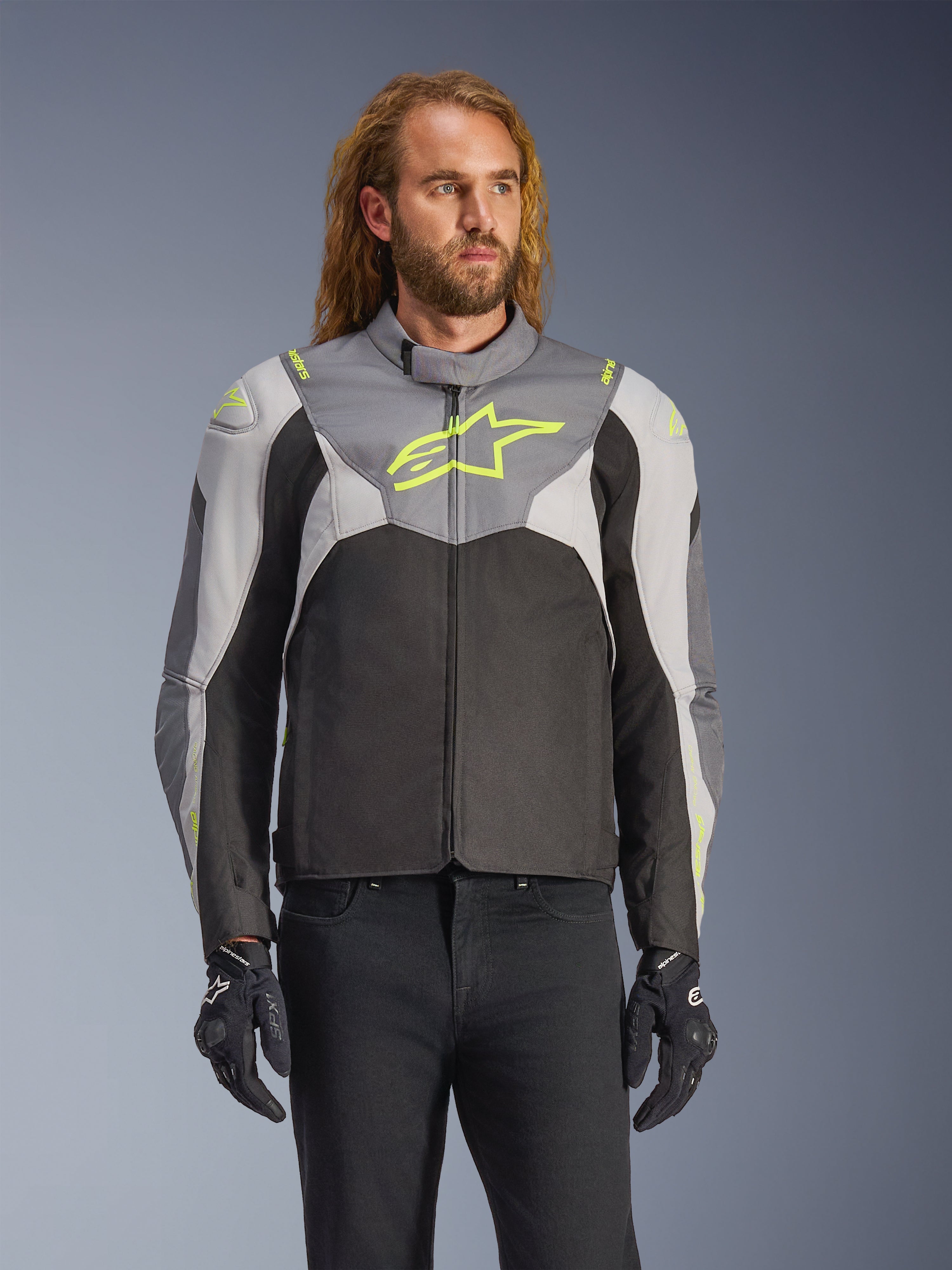 Alpinestars T-Jaws V4 Waterproof Jacke, getragen von einem männlichen Model, Schwarz, Eisgrau und Dunkelgrau mit neongelben Akzenten, markantes gelbes Stern-Logo auf der Brust, kombiniert mit schwarzer Hose und Motorradhandschuhen, Textil-Motorradjacke in Sport-Passform