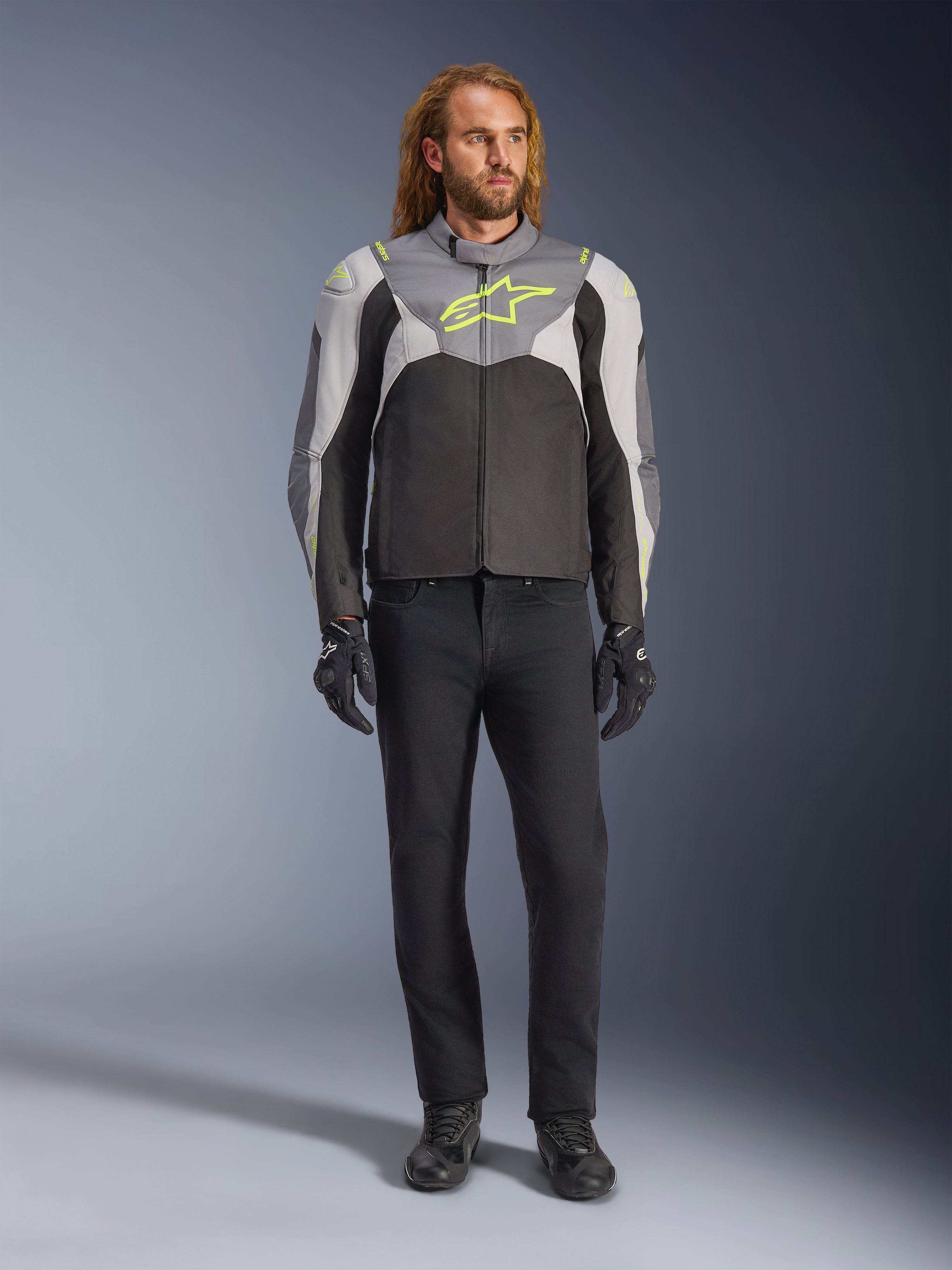 Alpinestars T-Jaws V4 Waterproof Jacke in Black Ice Gray Dark Gray mit neongelben Akzenten, getragen von einem männlichen Model, sportliche Silhouette, technische Textilkonstruktion, markantes Brustlogo, kombiniert mit schwarzer Motorradhose und Handschuhen