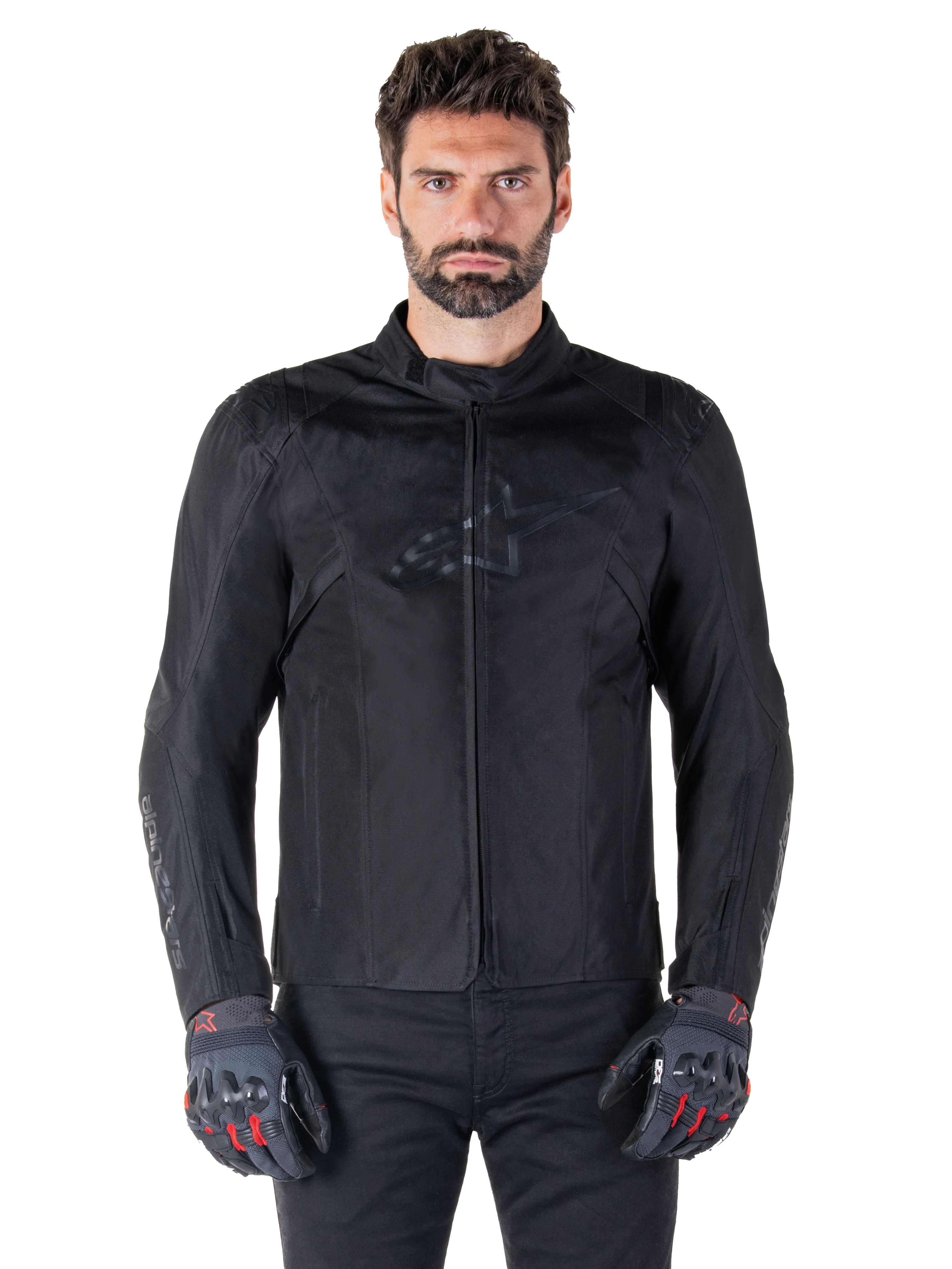 Alpinestars T-SPS V2 Waterproof Jacket, schwarze Motorrad-Textiljacke, getragen von einem Fahrer mit schwarz-roten Handschuhen, hoher Kragen, Reißverschluss vorne, dezentes Brustlogo, wasserdichter Schutz für sportliches und urbanes Fahren