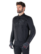 Model trägt Alpinestars T-SPS V2 Wasserdichte Jacke, Schwarz, kombiniert mit einer schwarzen Hose und grauen Motorradhandschuhen mit roten Akzenten, mit dezentem schwarzem Brustlogo und Branding am Ärmel, Textiljacke im Sportschnitt für den Straßeneinsatz.