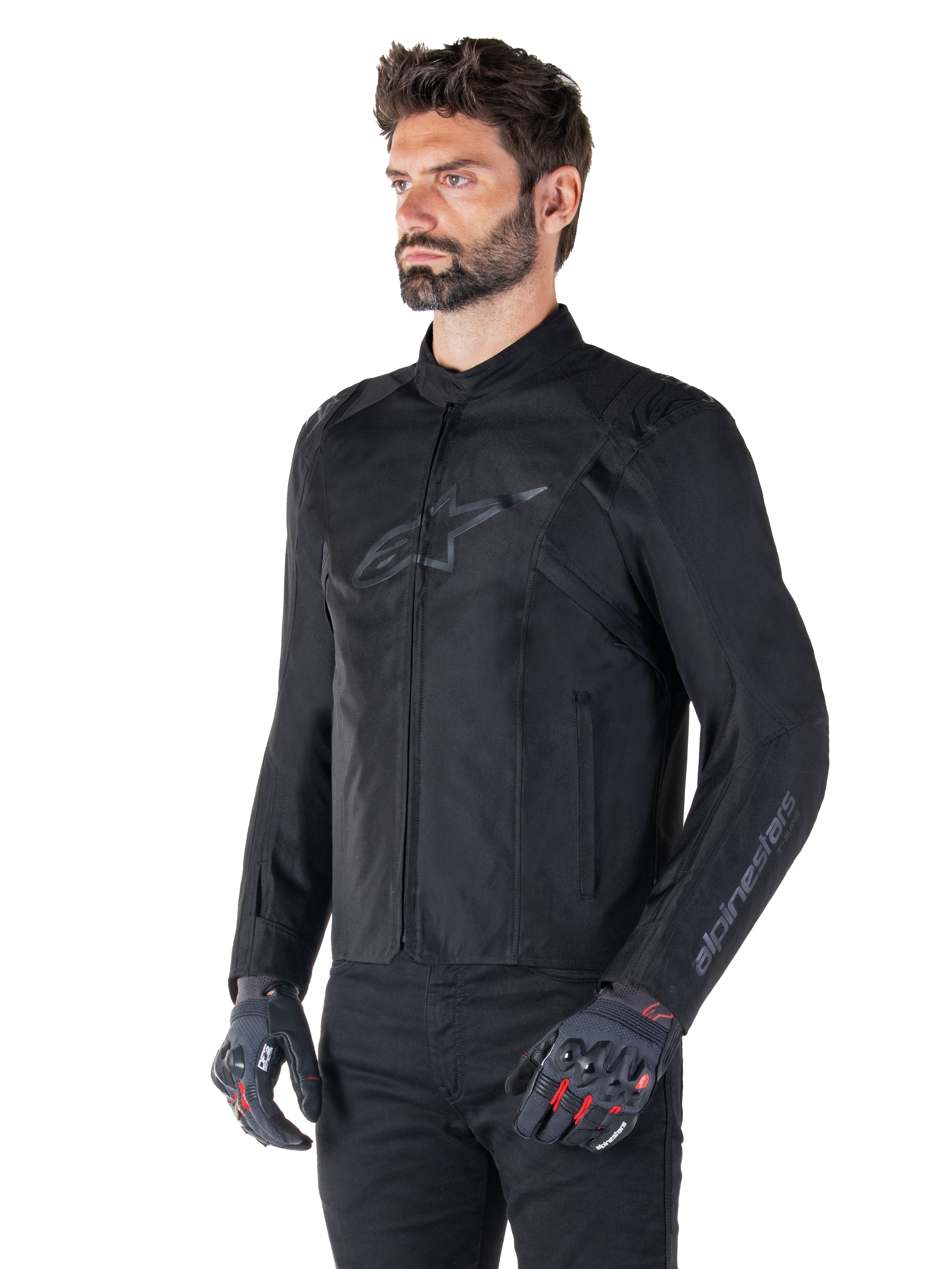 Model trägt Alpinestars T-SPS V2 Wasserdichte Jacke, Schwarz, kombiniert mit einer schwarzen Hose und grauen Motorradhandschuhen mit roten Akzenten, mit dezentem schwarzem Brustlogo und Branding am Ärmel, Textiljacke im Sportschnitt für den Straßeneinsatz.