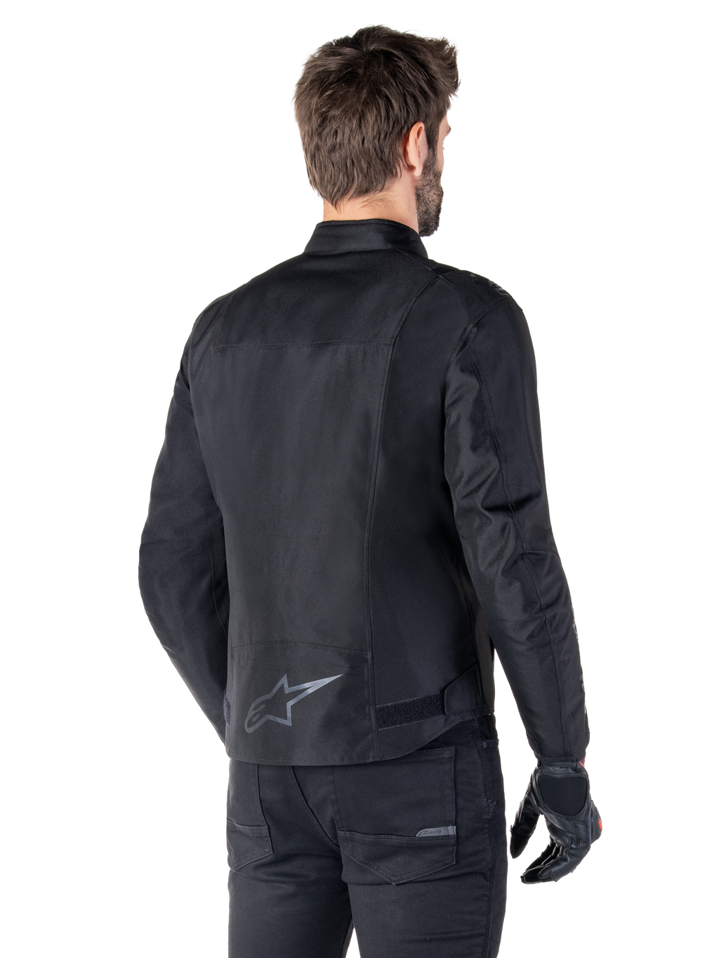 Alpinestars T-SPS V2 wasserdichte Jacke, Schwarz, Rückansicht an einem männlichen Model, mit reflektierendem Stern-Logo am unteren Rücken, verlängertem Rückenprofil und Taillenverstellriemen, kombiniert mit schwarzer Motorradhose und Handschuhen