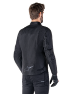 Alpinestars T-SPS V2 wasserdichte Jacke, Schwarz, Rückansicht an einem männlichen Model, mit reflektierendem Stern-Logo am unteren Rücken, verlängertem Rückenprofil und Taillenverstellriemen, kombiniert mit schwarzer Motorradhose und Handschuhen
