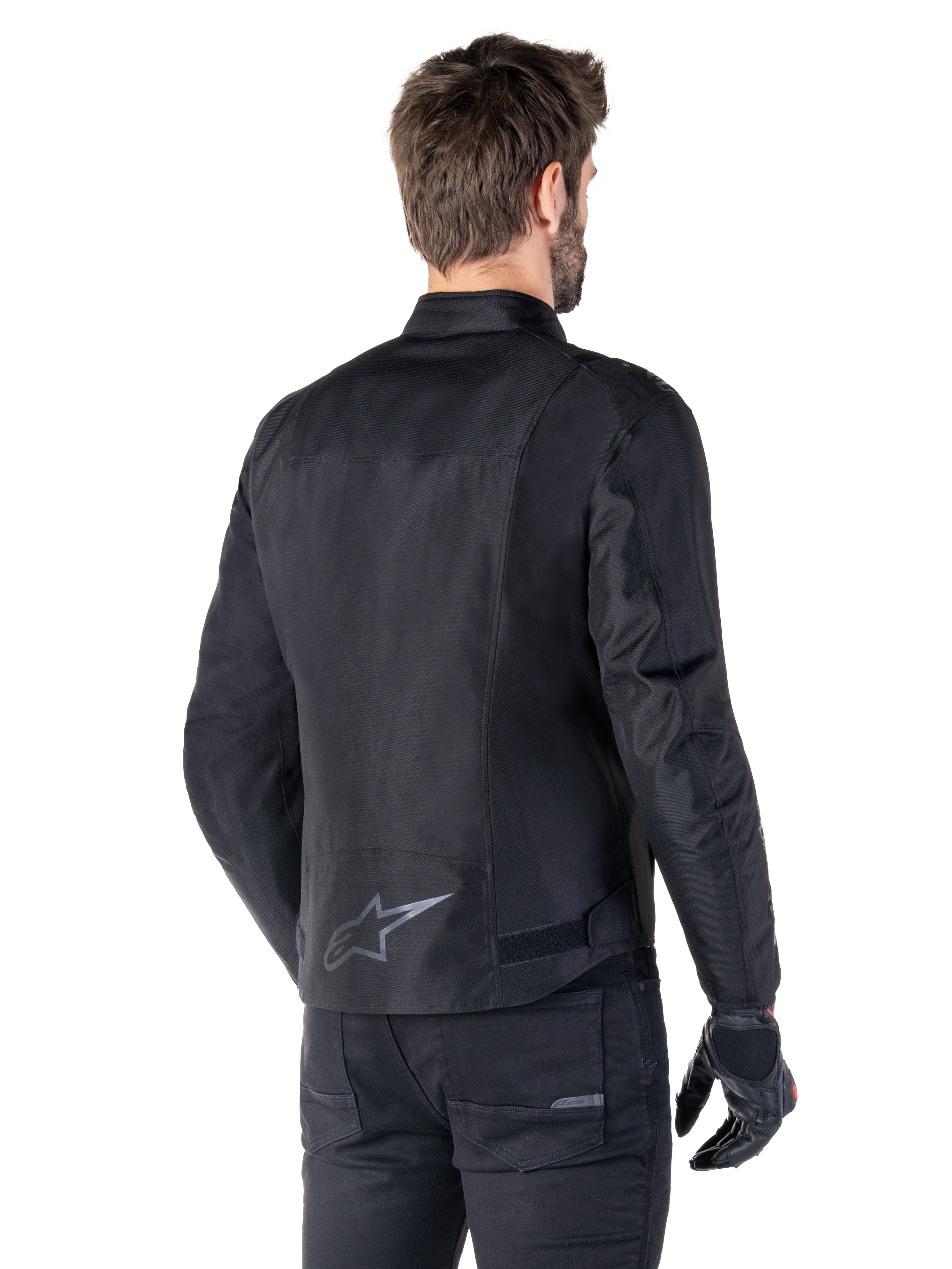 Alpinestars T-SPS V2 wasserdichte Jacke, Schwarz, Rückansicht an einem männlichen Model, mit reflektierendem Stern-Logo am unteren Rücken, verlängertem Rückenprofil und Taillenverstellriemen, kombiniert mit schwarzer Motorradhose und Handschuhen