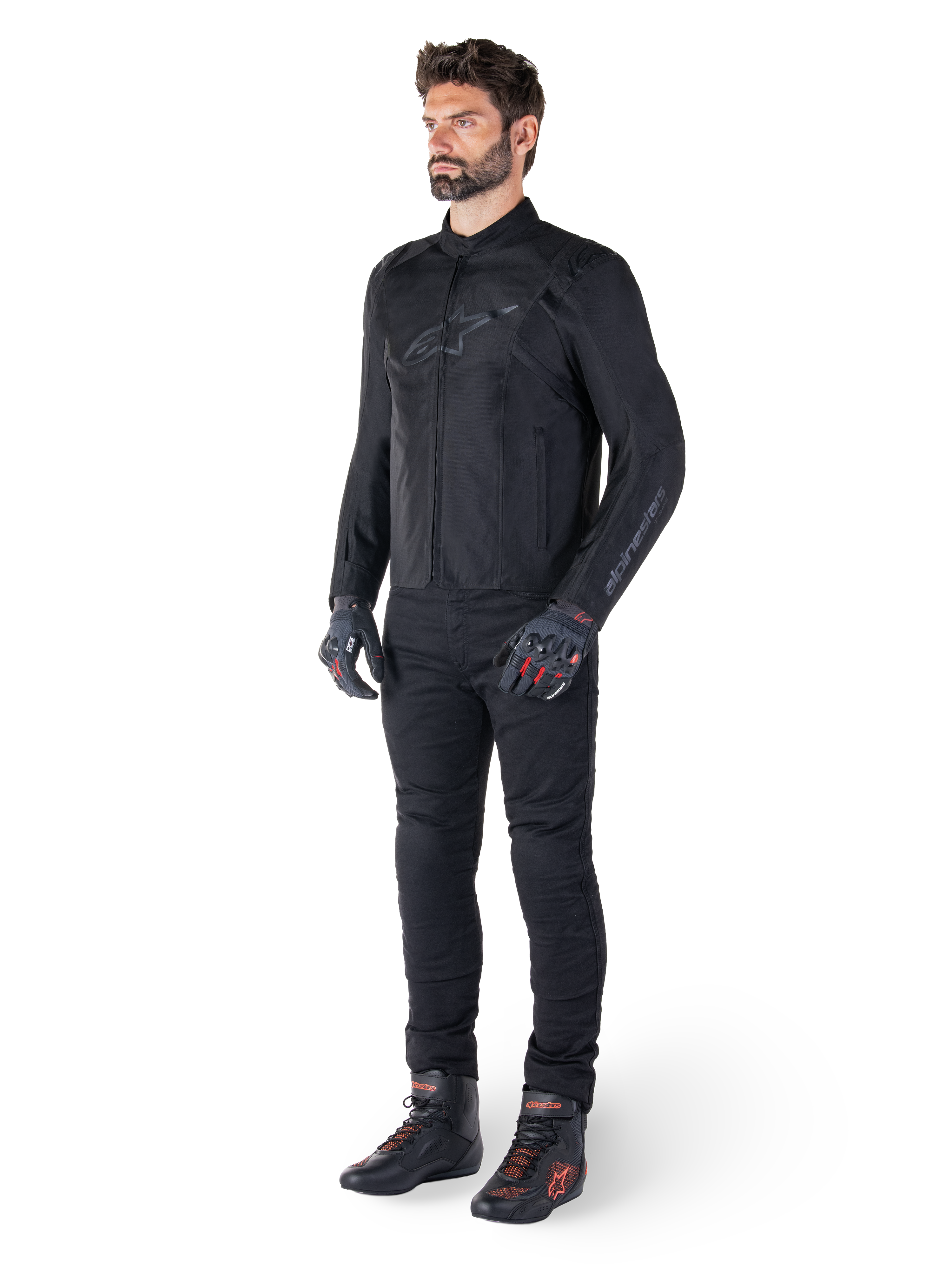 Alpinestars T-SPS V2 Waterproof Jacke, Schwarz, getragen von einem männlichen Model mit passender schwarzer Hose, schwarzen Handschuhen mit roten Akzenten und schwarzen Motorradschuhen, komplette Motorradausrüstung für Urban- und Sport-Fahrten, sauberer weißer Hintergrund