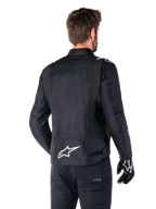 Alpinestars T-SPS V2 Wasserdichte Jacke, Schwarz und Weiß, Rückansicht eines männlichen Fahrers, der die Jacke trägt, weißes Alpinestars-Logo auf dem unteren Rücken, Textilkonstruktion im Sportschnitt, getragen mit schwarzer Motorradhose und Handschuhen