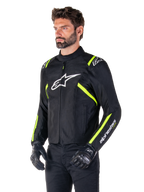 Männliches Model in der Alpinestars T-SPS V2 Waterproof Jacke, Schwarz mit weißen und gelben Fluo-Akzenten, mit einem weißen Astars-Logo auf der Brust und fluoreszierenden gelben Streifen an den Schultern und Ärmeln, kombiniert mit einer schwarzen Hose und Motorradhandschuhen, sportlich geschnittene Textil-Motorradjacke.