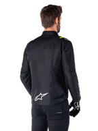 Alpinestars T-SPS V2 Waterproof Jacke, Schwarz Weiß Gelb Fluo, Rückansicht eines männlichen Models, das die Textil-Motorradjacke trägt, mit einem weißen Logo am unteren Rücken und gelben Fluo-Akzenten an den Schultern, kombiniert mit schwarzer Hose und Handschuhen.