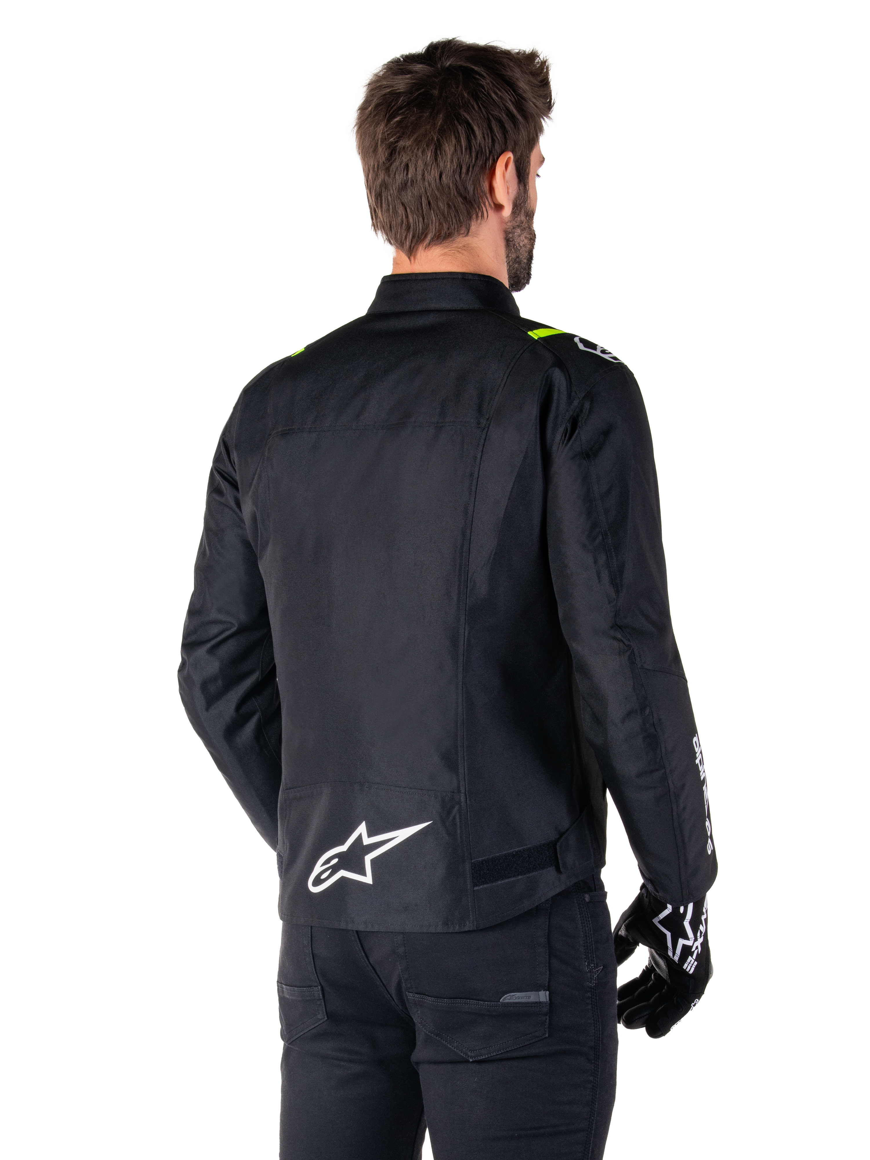 Alpinestars T-SPS V2 Waterproof Jacke, Schwarz Weiß Gelb Fluo, Rückansicht eines männlichen Models, das die Textil-Motorradjacke trägt, mit einem weißen Logo am unteren Rücken und gelben Fluo-Akzenten an den Schultern, kombiniert mit schwarzer Hose und Handschuhen.