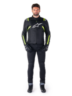 Alpinestars T-SPS V2 Waterproof Jacke, Schwarz, Weiß und Gelb Fluo, getragen von einem Model mit schwarzer Motorradhose und Handschuhen, mit weißem Alpinestars-Logo auf der Brust und fluoreszierenden Akzenten an Schultern und Ärmeln, sportlich geschnittene Motorradbekleidung