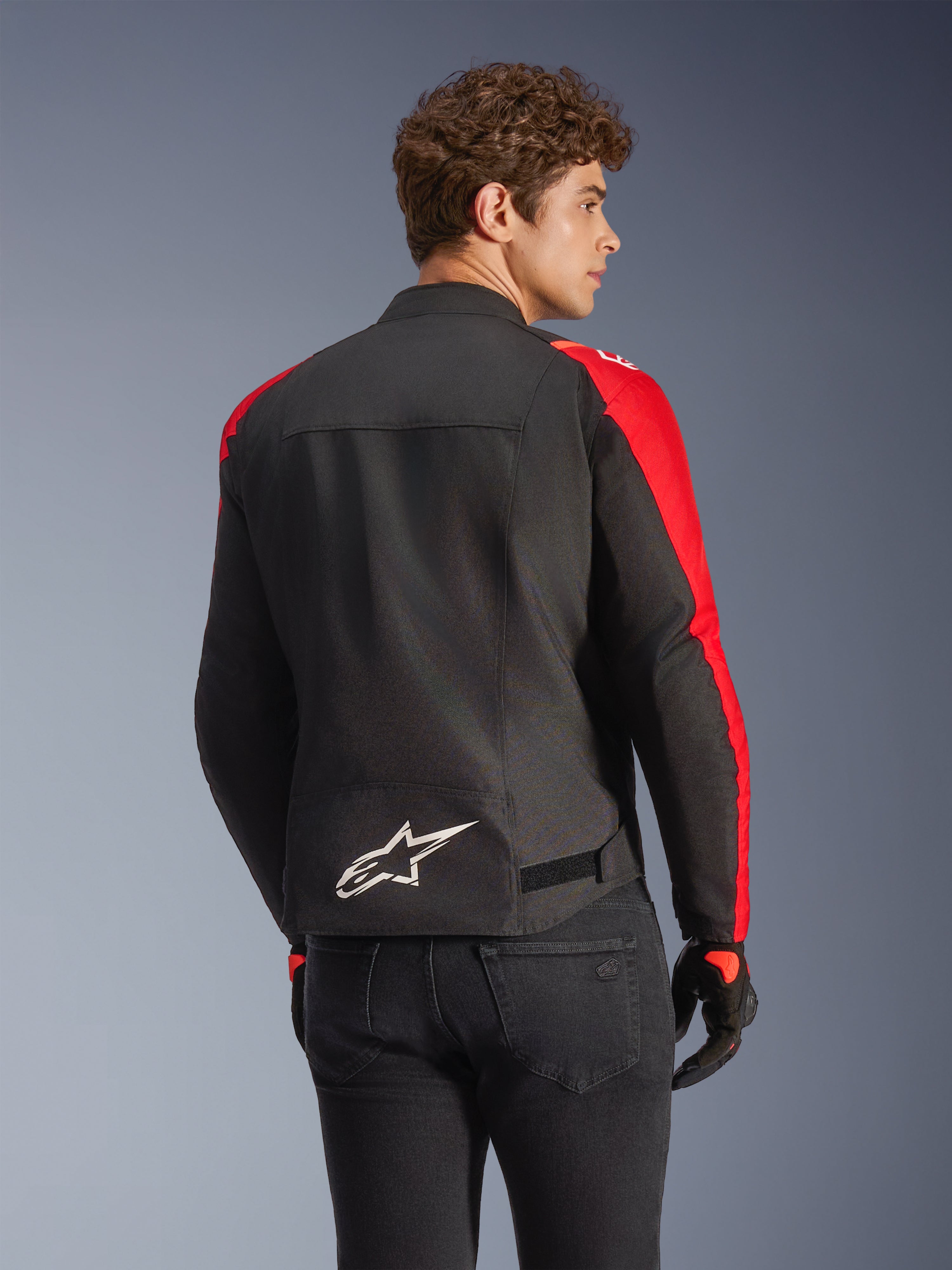 Alpinestars T-SPS V2 Wasserdichte Jacke in Schwarz und Rot, Rückansicht an einem Modell, mit weißem Alpinestars-Logo auf dem unteren Rücken, leuchtend roten Ärmeleinsätzen und einer sportlichen Silhouette, getragen mit schwarzen Handschuhen und Jeans.