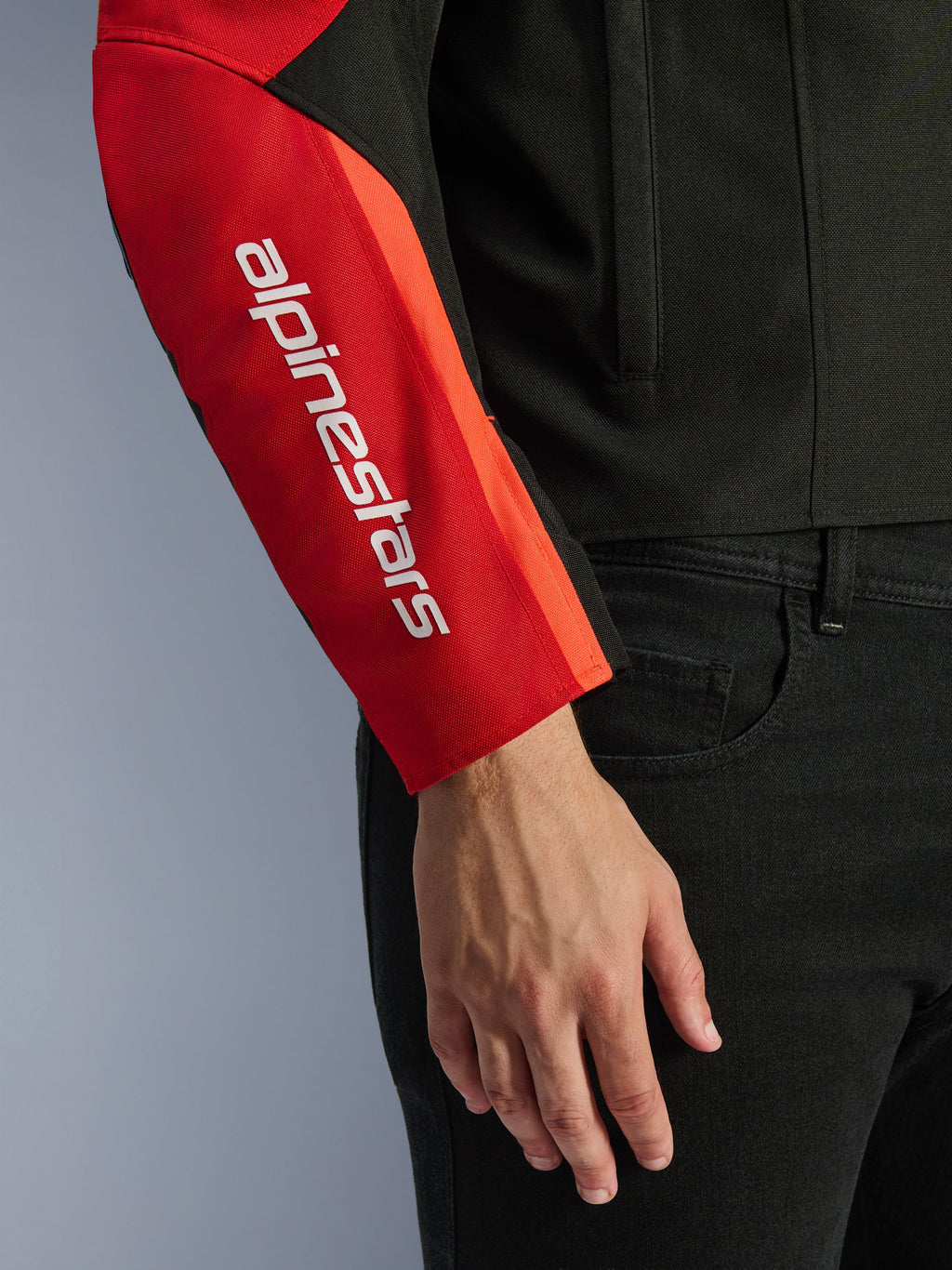 Alpinestars T-SPS V2 Wasserdichte Jacke, Motorradjacke, Schwarz und Rot, Nahaufnahme des Ärmels mit einem weißen vertikalen Markenlogo auf einem roten Einsatz, strapazierfähige Textilkonstruktion mit sportlich inspiriertem Design.