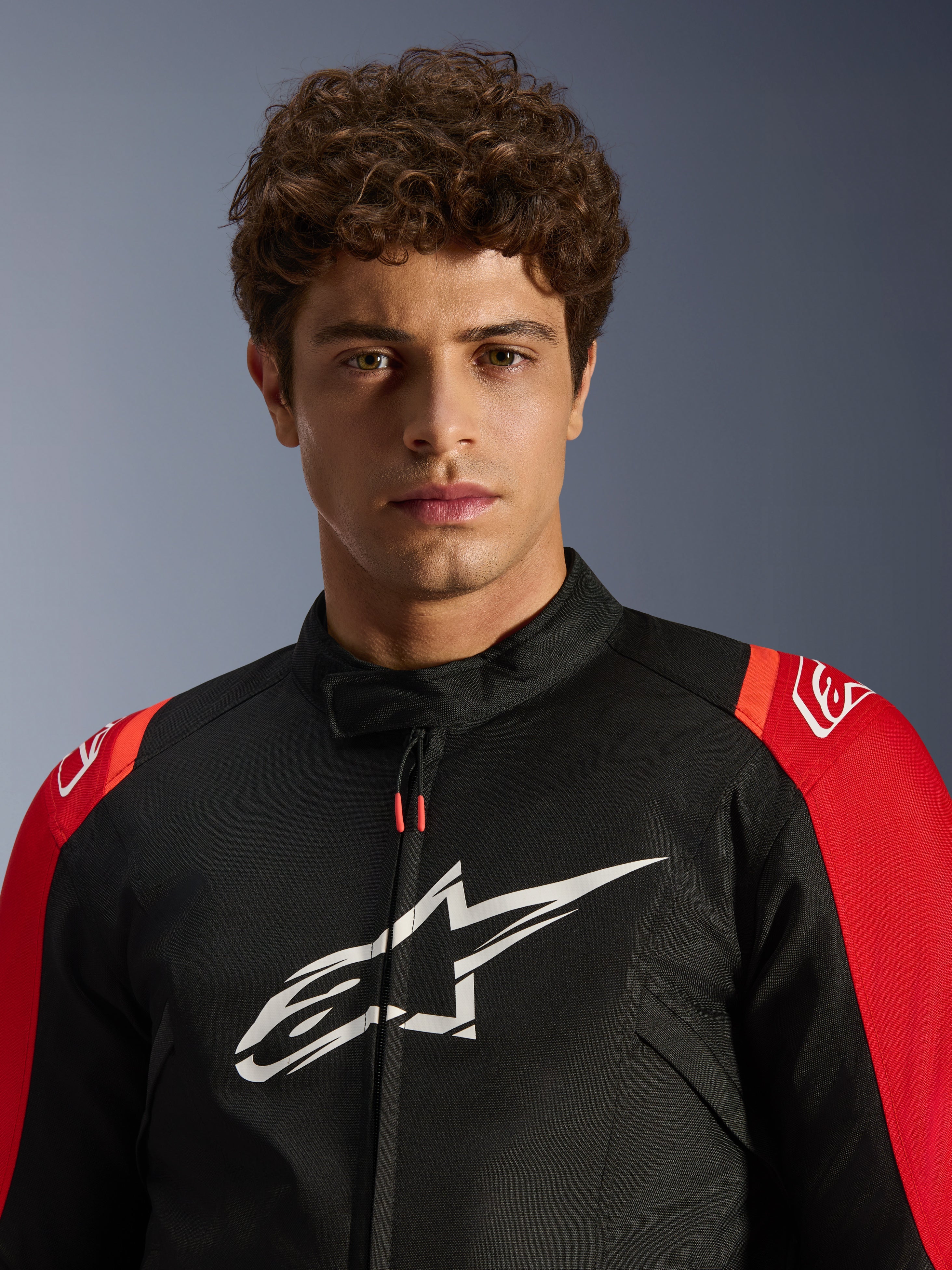 Männliches Model in der Alpinestars T-SPS V2 Waterproof Jacke, Schwarz mit roten Akzenten und weißem Logo, Nahaufnahme Porträtansicht, sportliche Textil-Motorradjacke
