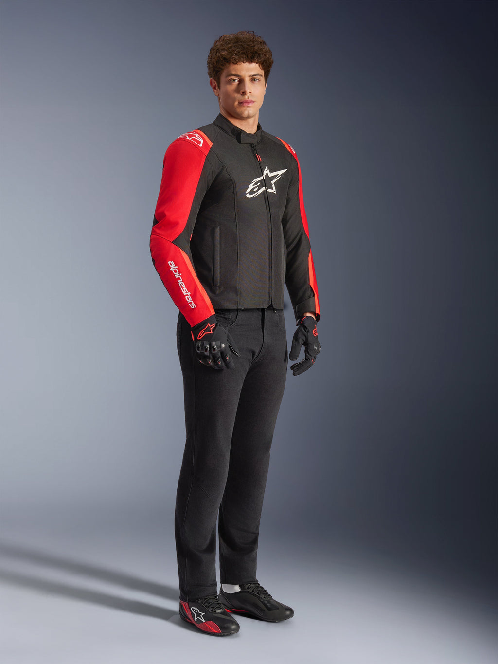 Männliches Model in der Alpinestars T-SPS V2 Waterproof Jacke in Schwarz und Rot Fluo, mit auffällig roten Ärmeln samt weißem Branding und einem großen weißen Logo auf der Brust, kombiniert mit schwarzer Motorradhose, Motorradhandschuhen und flachen Motorradschuhen, Studioaufnahme in voller Länge.