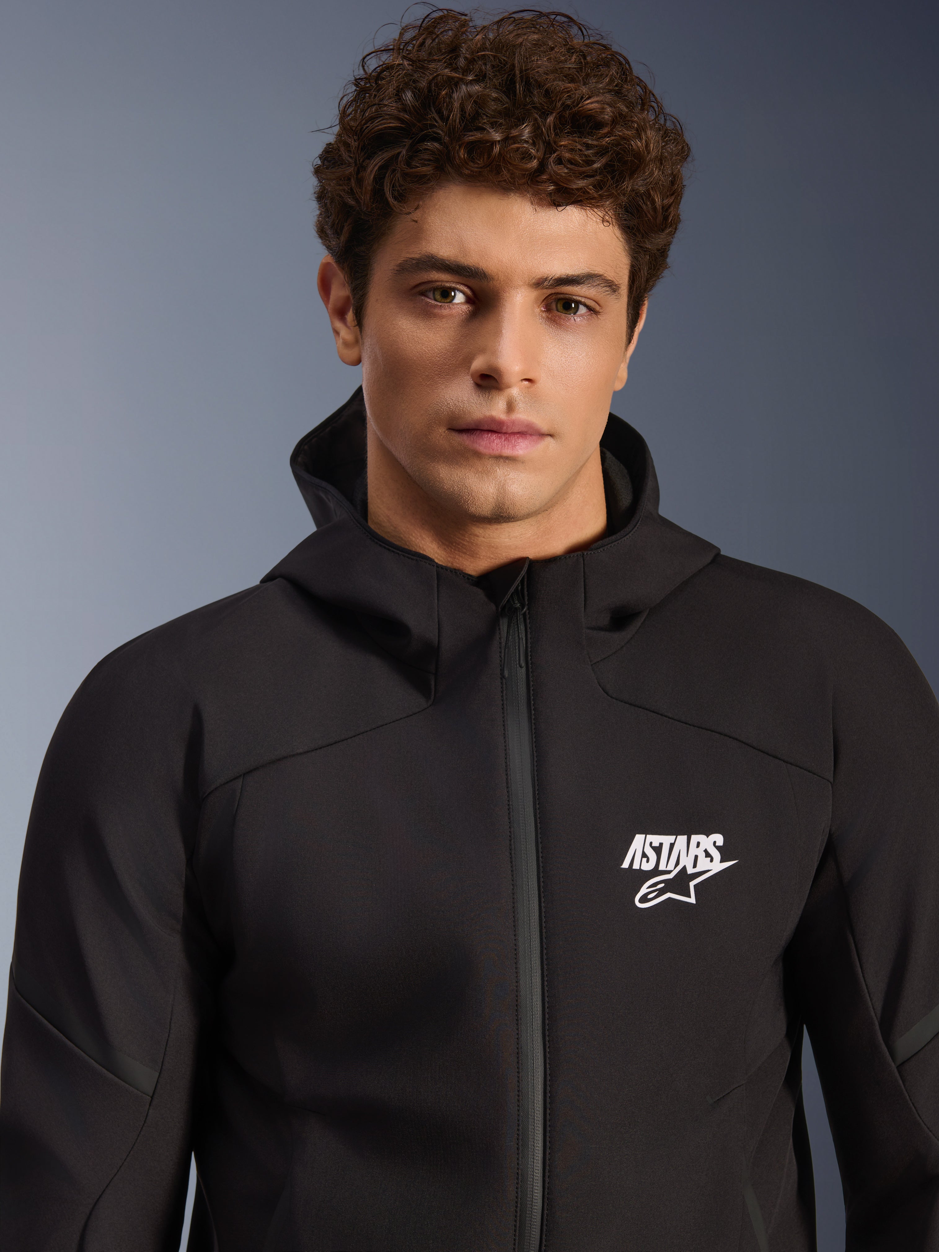 Alpinestars Aeroshell Wasserdichte Jacke, schwarzer Motorrad-Softshell-Hoodie für Herren, weißes ASTARS-Logo, wasserdichtes Textilmaterial, modernes Studioporträt.