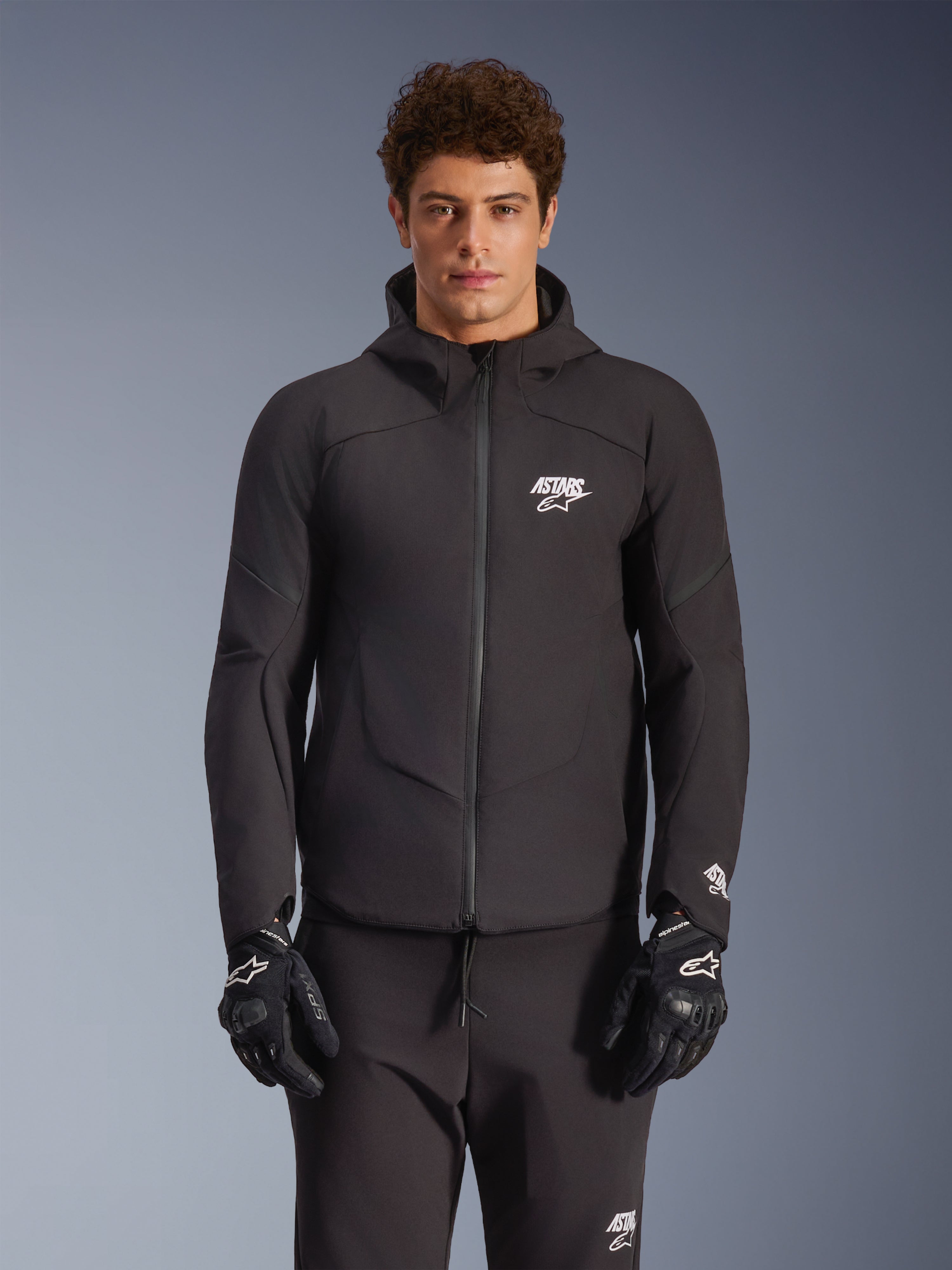 Alpinestars Aeroshell Wasserdichte Jacke, Urban-Motorradjacke für Herren, Schwarz, Textil-Softshell mit integrierter Kapuze, weißes Logo auf der Brust, getragen von einem Model mit schwarzen Handschuhen und Hose.
