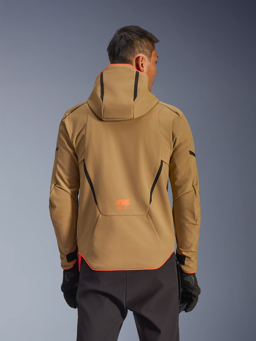 Alpinestars Aeroshell WP Jacke, wasserdichte Jacke, Dark Khaki und Orange Fluo, Rückansicht zeigt Hoodie-Design mit orangefarbenem Besatz an Kapuze und Saum, orangefarbenes Logo am unteren Rücken, getragen mit schwarzen Handschuhen, Crossover-Sport-Softshell-Motorradausrüstung