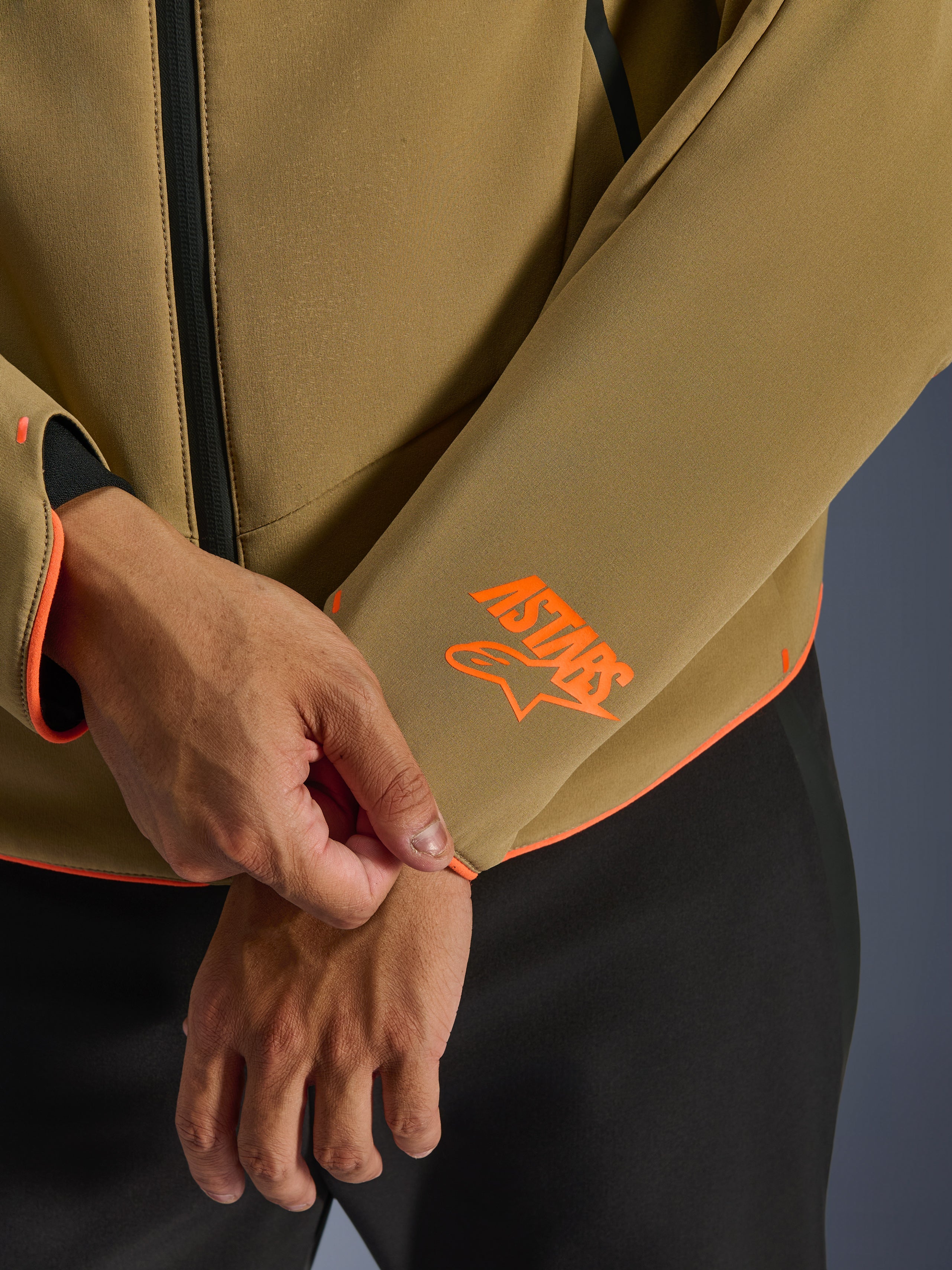 Alpinestars Aeroshell Waterproof Jacke, Dunkelkhaki mit Orange Fluo Akzenten, Nahaufnahme des Ärmelbündchens und des unteren Saums mit leuchtend orangefarbenem Astars-Logo, neon-orangener Paspelierung, wasserdichter Frontreißverschluss, sportliche Softshell-Konstruktion