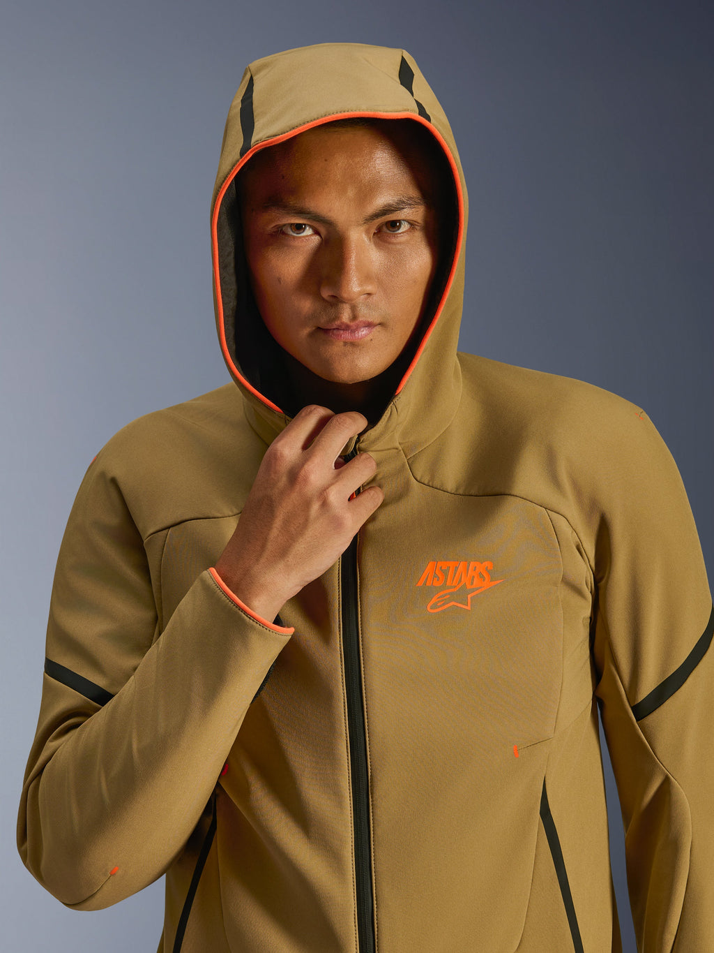 Alpinestars Aeroshell wasserdichte Jacke, Dark Khaki Black Orange Fluo, Modell trägt die Jacke mit aufgesetzter Kapuze, mit leuchtend orangefarbenen Fluo-Besätzen und einem orangefarbenen ASTARS-Logo, elegante schwarze Akzente, moderne wasserdichte Sport-Softshell