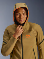 Alpinestars Aeroshell wasserdichte Jacke, Dark Khaki Black Orange Fluo, Modell trägt die Jacke mit aufgesetzter Kapuze, mit leuchtend orangefarbenen Fluo-Besätzen und einem orangefarbenen ASTARS-Logo, elegante schwarze Akzente, moderne wasserdichte Sport-Softshell