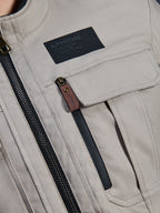 Alpinestars Borrego Drystar® Jacke, Warm Gray, Nahaufnahme des Brustbereichs mit einem schwarzen Alpinestars-Markenaufnäher, Brusttasche mit Klappe und einem vertikalen Reißverschluss mit brauner Lederlasche, robuste graue Textil-Canvas-Konstruktion.