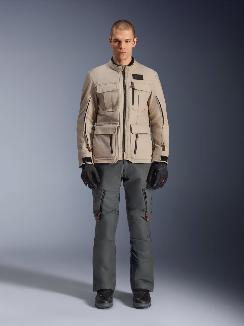 Alpinestars Borrego Drystar® Jacke, Adventure-Motorradjacke, Warm Gray, getragen von einem Modell mit dunkelgrauer Fahrhose und schwarzen Handschuhen, mit mehreren Cargo-Taschen und einer strapazierfähigen Textilkonstruktion im klassischen gewachsten Canvas-Look.
