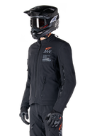 Alpinestars AMT-8 Stretch Drystar® XF Jacke, Schwarz, getragen von einem Fahrer mit passender Adventure-Hose und Handschuhen, mit einem schwarzen Motocross-Helm und einer Schutzbrille, Belüftungsöffnungen mit Reißverschluss an Brust und Armen, Alpinestars Motion Technology-Branding mit einem orangefarbenen Logo, Premium-Adventure-Touring-Ausrüstung.