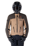 Alpinestars AMT-8 Stretch Drystar XF Jacke, Motorrad-Adventure-Touringjacke, Dunkelkhaki und Dunkelbraun, mit orangefarbenem Astars-Logo und schwarzen Reißverschlüssen, getragen mit schwarzem Adventure-Helm und Handschuhen, komplette Fahrausrüstung