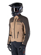 Alpinestars AMT-8 Stretch Drystar® XF Jacke, Adventure-Motorradjacke, Dunkelbraun und Dunkles Khaki, getragen mit einem schwarzen Offroad-Helm und Goggles, mit Alpinestars Motion Technology Branding und Belüftungsöffnungen mit Reißverschluss.