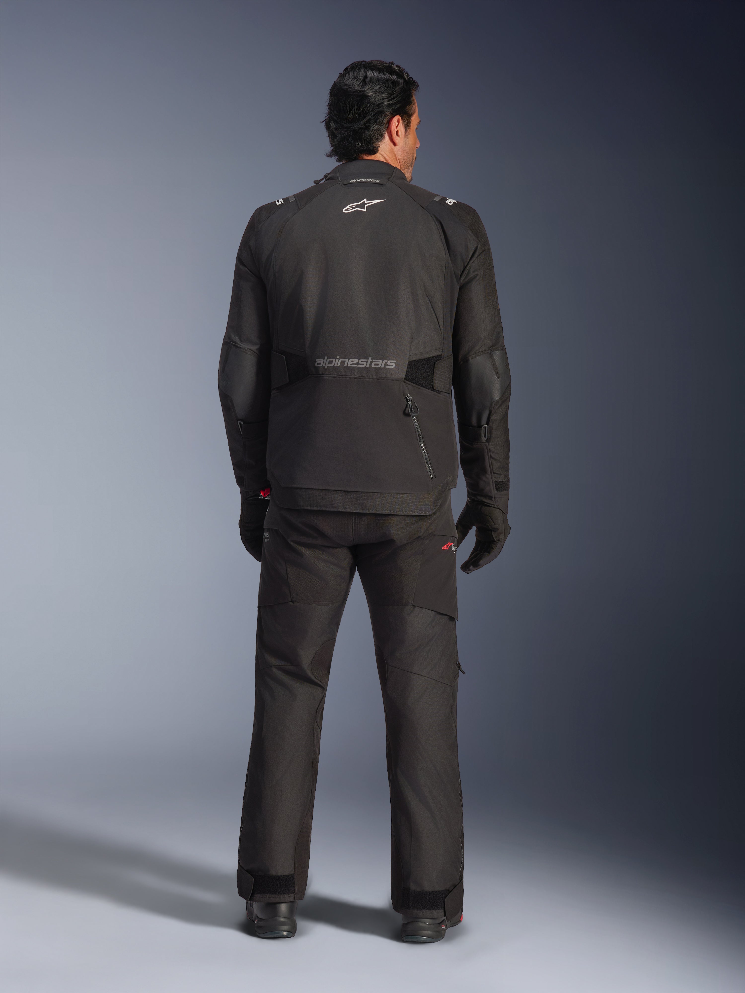 Rückansicht eines männlichen Fahrers in der Alpinestars Andes V4 Drystar® Jacke in Schwarz, kombiniert mit passender Motorradhose und Handschuhen, mit Alpinestars-Logo und -Schriftzug auf dem Rücken, technische Allwetter-Adventure-Ausrüstung