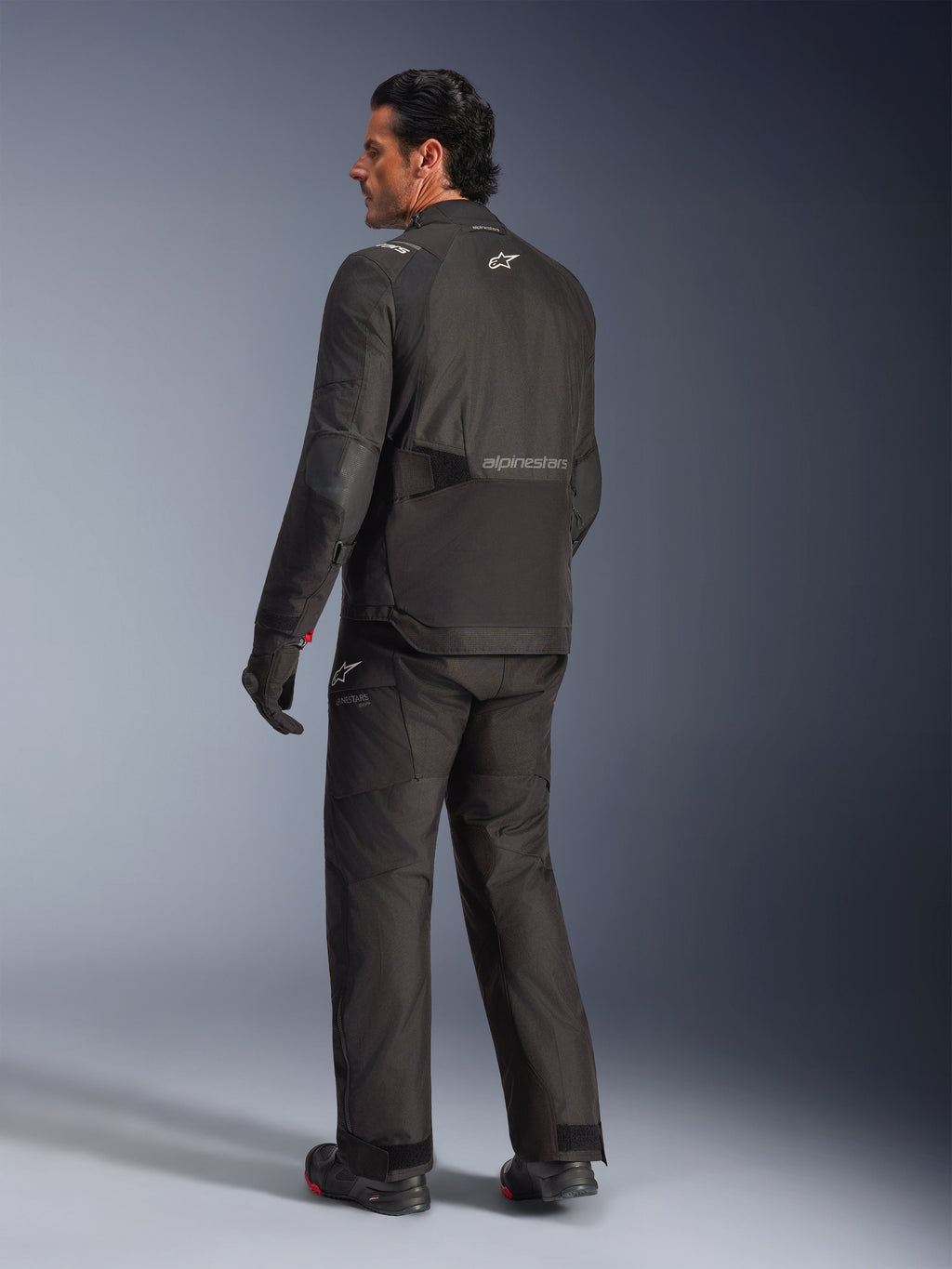 Alpinestars Andes V4 Drystar® Jacke, Schwarz, Rückansicht eines Fahrers in kompletter Motorradausrüstung, Textil-Adventure-Jacke mit Alpinestars-Branding auf dem Rücken, getragen mit passender schwarzer Hose und Stiefeln, Allwetter-Straßen- und Adventure-Fahrstil