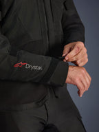 Alpinestars Andes V4 Drystar® Jacke, schwarze Textil-Motorradjacke, Nahaufnahme des Ärmels mit rotem Alpinestars-Logo und weißem Drystar®-Branding, Fahrer passt den Klettverschluss am Handgelenk für einen sicheren Sitz an, funktionales Detail für Adventure-Fahrten