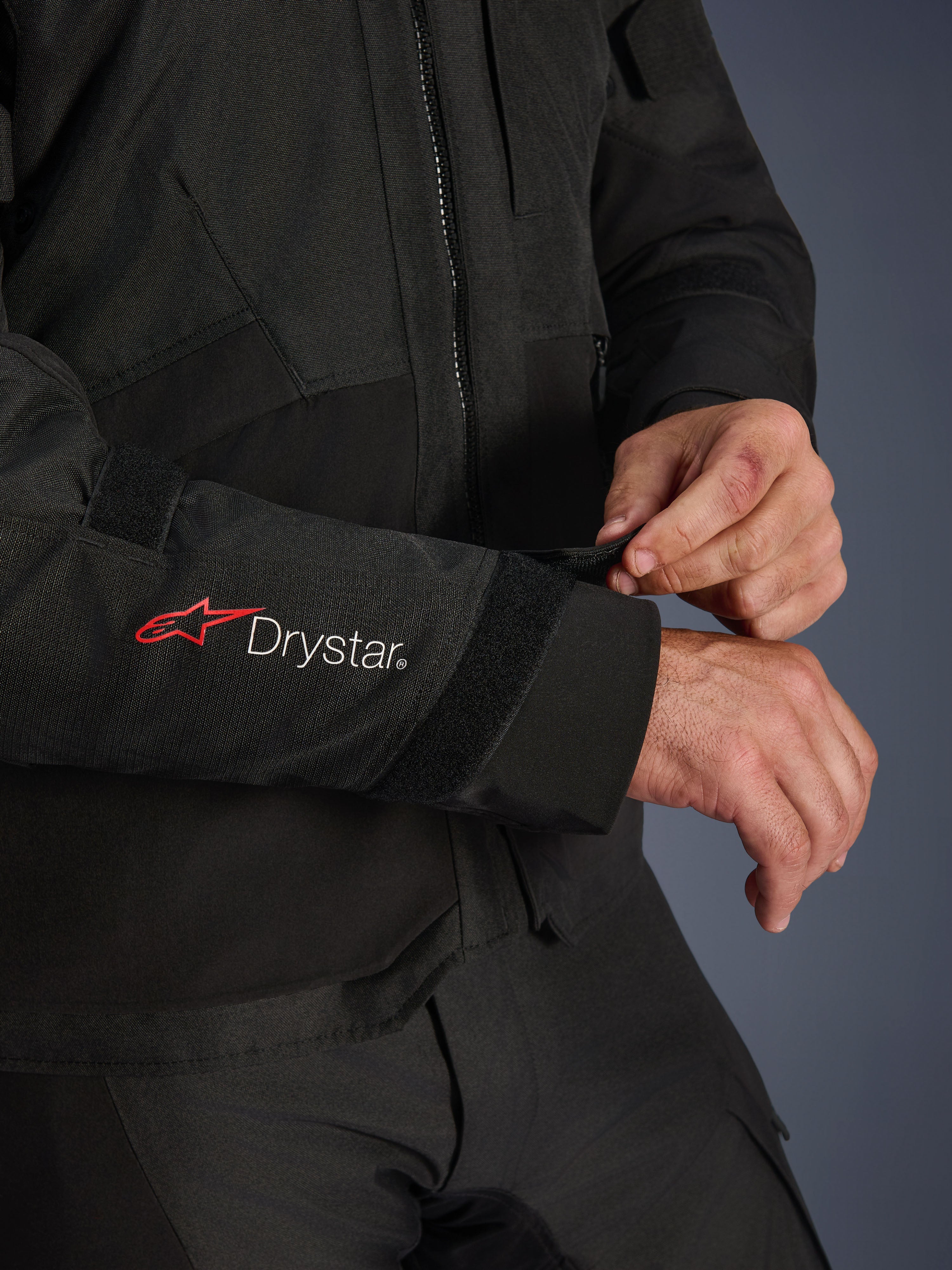 Alpinestars Andes V4 Drystar® Jacke, schwarze Textil-Motorradjacke, Nahaufnahme des Ärmels mit rotem Alpinestars-Logo und weißem Drystar®-Branding, Fahrer passt den Klettverschluss am Handgelenk für einen sicheren Sitz an, funktionales Detail für Adventure-Fahrten