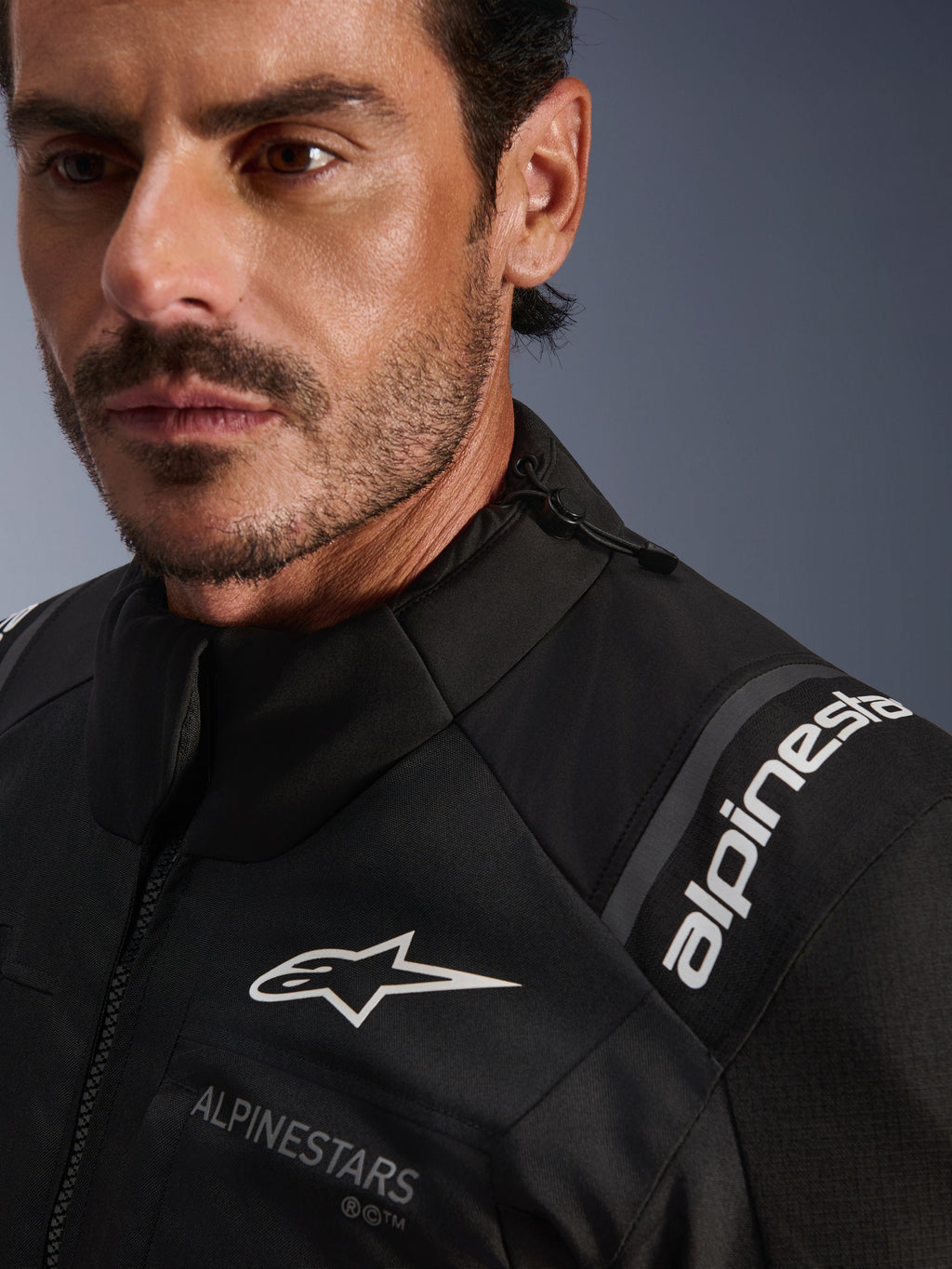 Alpinestars Andes V4 Drystar® Jacke, schwarze Textil-Motorradjacke, Nahaufnahme eines Mannes, mit weißen Alpinestars-Logos auf Brust und Schulter, verstellbarem Kragen-Kordelzug und leistungsstarkem Adventure-Touring-Design.