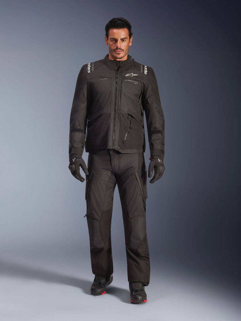 Männliches Model in der Alpinestars Andes V4 Drystar® Jacke in Schwarz, kombiniert mit passender Adventure-Motorradhose, schwarzen Handschuhen und schwarzen Stiefeln, Ganzkörper-Studioaufnahme technischer Allwetter-Tourenbekleidung mit Alpinestars-Branding.