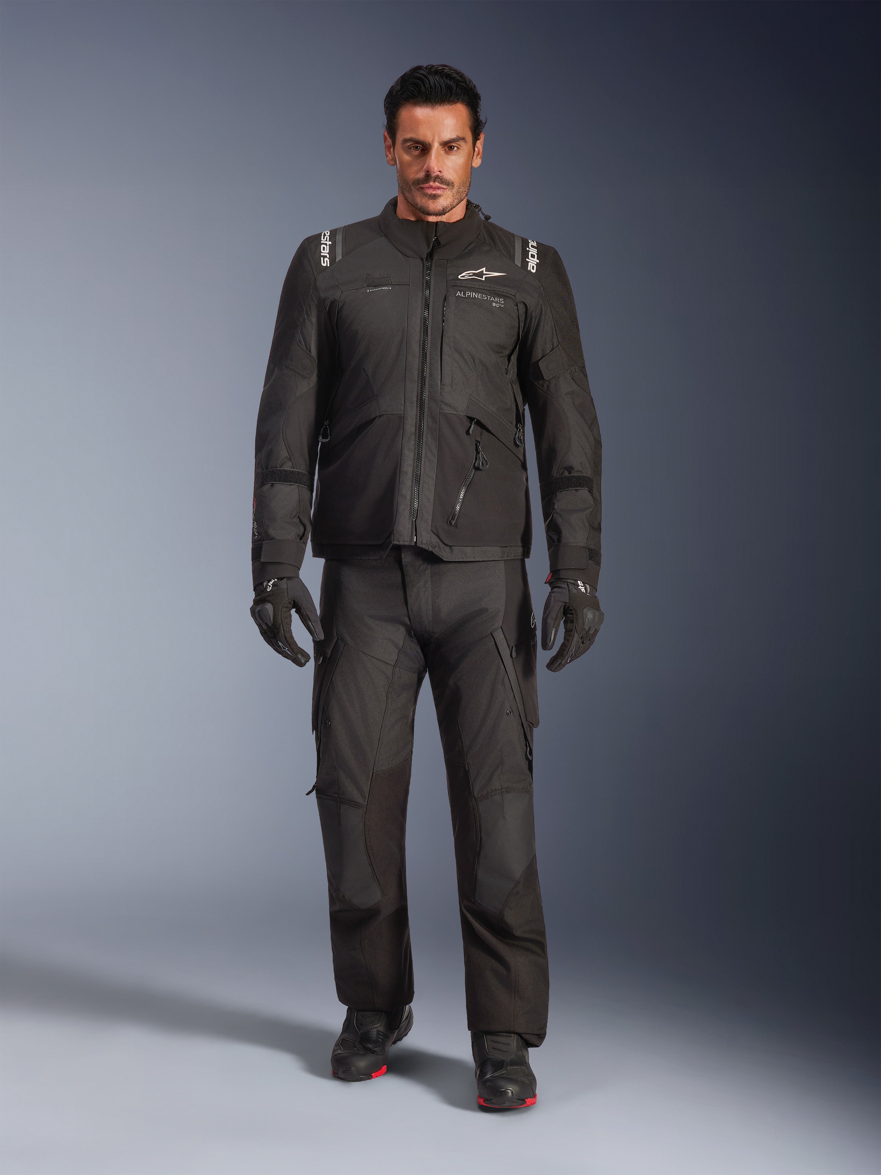 Männliches Model in der Alpinestars Andes V4 Drystar® Jacke in Schwarz, kombiniert mit passender Adventure-Motorradhose, schwarzen Handschuhen und schwarzen Stiefeln, Ganzkörper-Studioaufnahme technischer Allwetter-Tourenbekleidung mit Alpinestars-Branding.