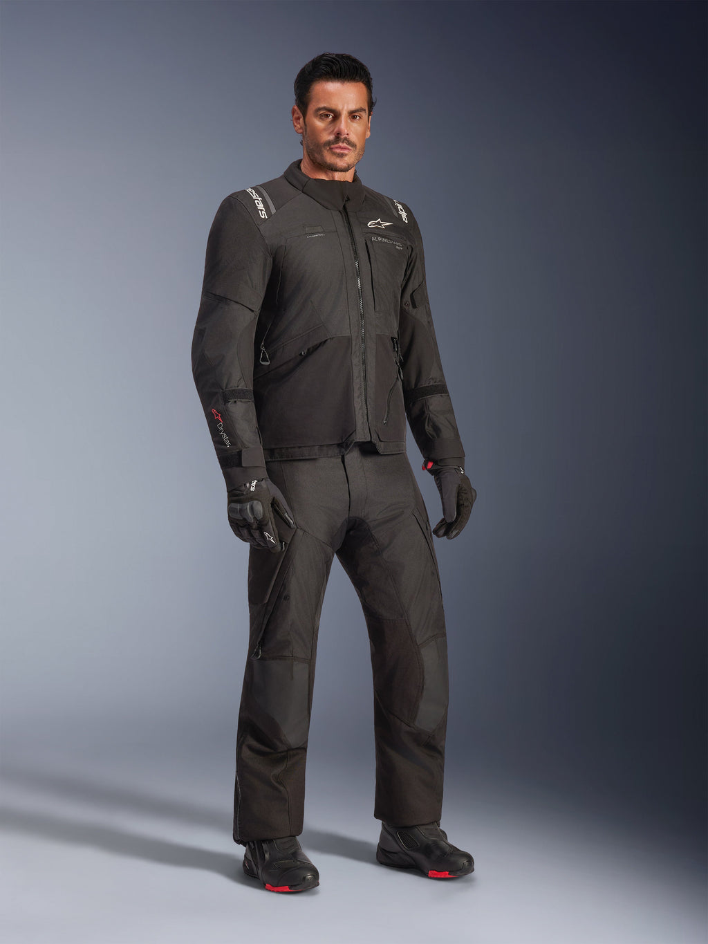 Alpinestars Andes V4 Drystar® Jacke, schwarzes Motorradbekleidungs-Set, getragen von einem Modell, Adventure-Touring-Textiljacke und -hose, schwarze Handschuhe und Stiefel, Studioaufnahme, modernes Design für das Fahren bei jedem Wetter