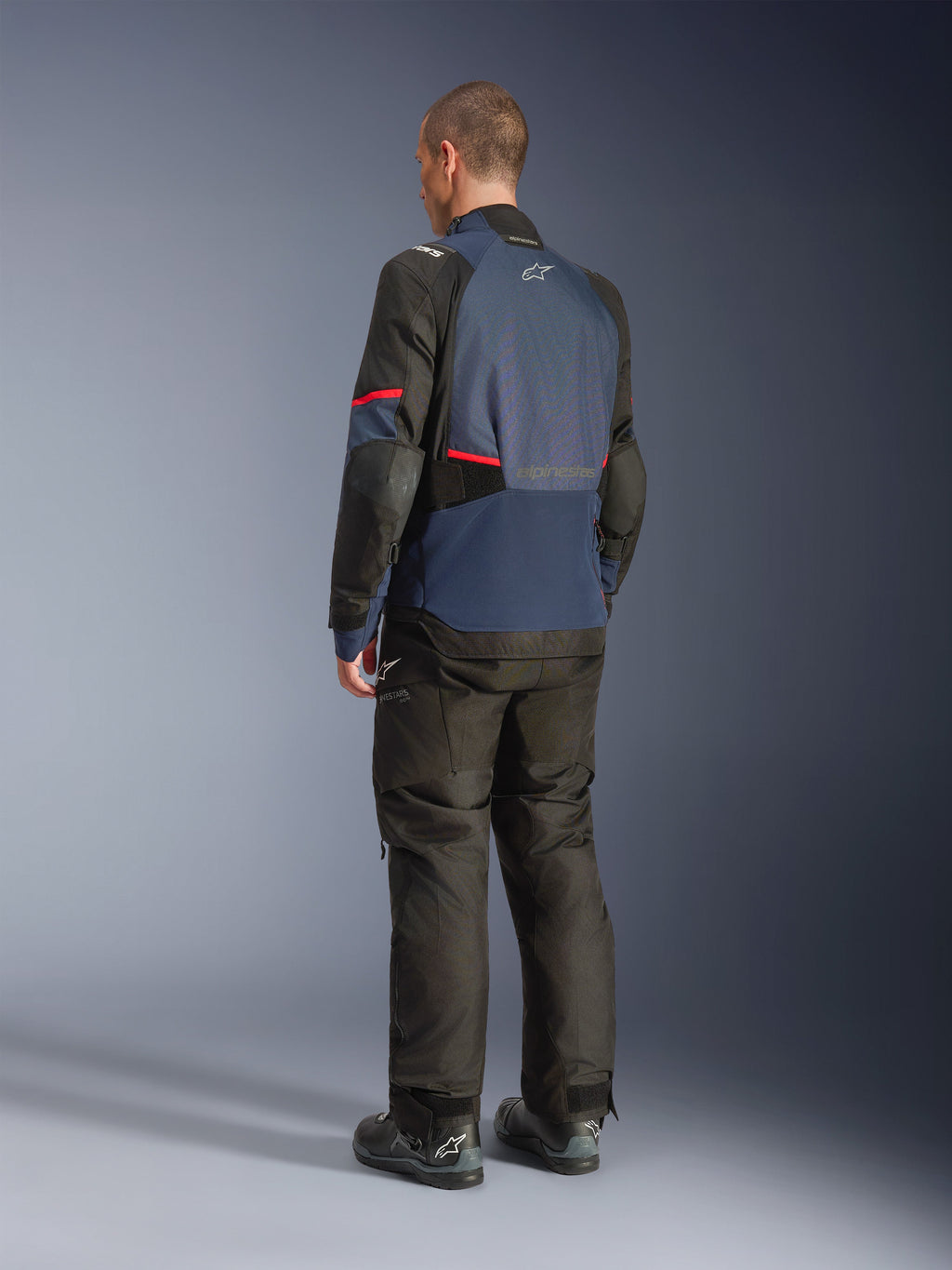 Alpinestars Andes V4 Drystar® Jacke, Night Navy und Mood Indigo mit schwarzen Einsätzen und roten Akzenten, Drei-Viertel-Rückansicht, getragen mit passender schwarzer Adventure-Fahrhose und Stiefeln, technische Textilkonstruktion mit Alpinestars-Logo auf dem oberen Rücken, verlängerter Rückensaum für verbesserte Abdeckung.