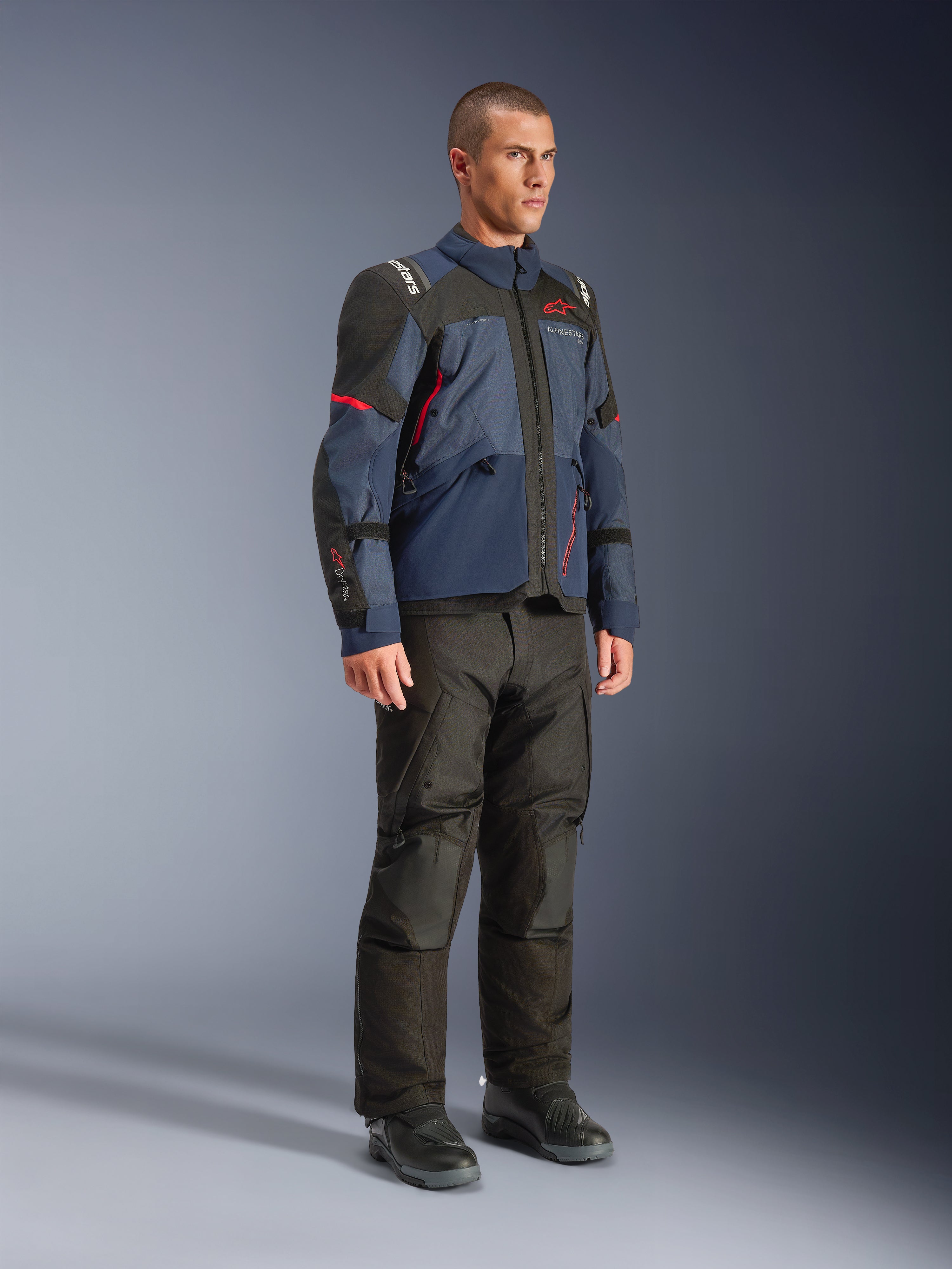 Männliches Modell trägt die Alpinestars Andes V4 Drystar® Jacke in Night Navy und Mood Indigo mit schwarzen Einsätzen und roten Akzenten, kombiniert mit einer schwarzen Motorradhose und Stiefeln, technische Adventure-Touring-Ausrüstung in einer Studioumgebung.