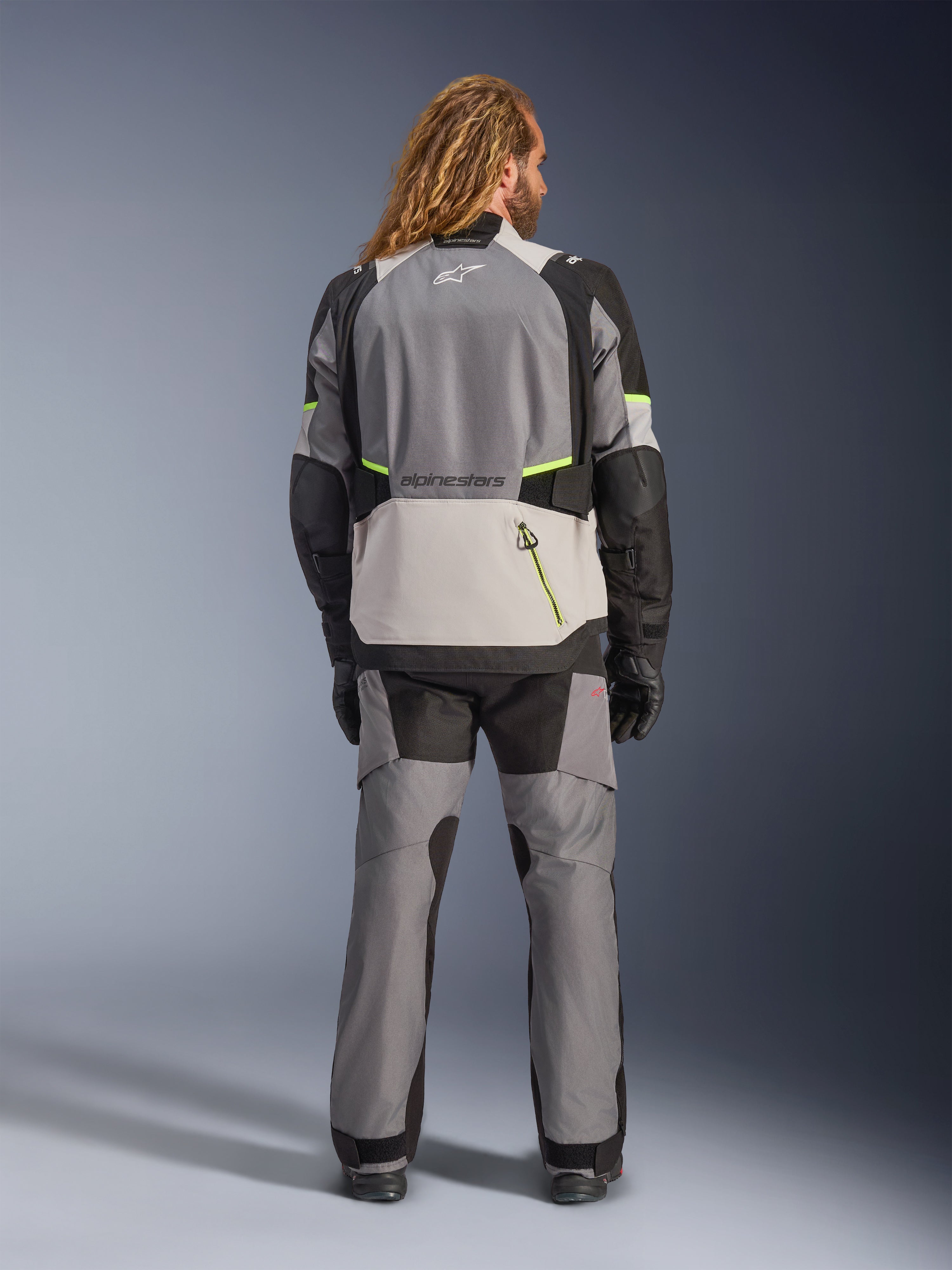 Alpinestars Andes V4 Drystar® Jacke, Adventure-Motorradjacke, Dunkelgrau und Eisgrau mit schwarzen und neongelben Akzenten, Rückansicht getragen mit passender Hose und schwarzen Handschuhen, große Rückentasche mit gelbem Reißverschluss, verstärkte Schulter- und Ellbogenpartien