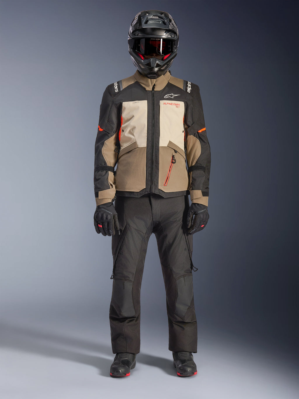 Alpinestars Andes V4 Drystar® Jacke, Walnut Mastic Black, Adventure-Tourenmotorradjacke mit hellbraunen und cremefarbenen Einsätzen und orangefarbenen Akzenten, getragen von einem Fahrer mit schwarzem Adventure-Helm, schwarzer Motorradhose und schwarzen Stiefeln, Ganzkörper-Studioansicht