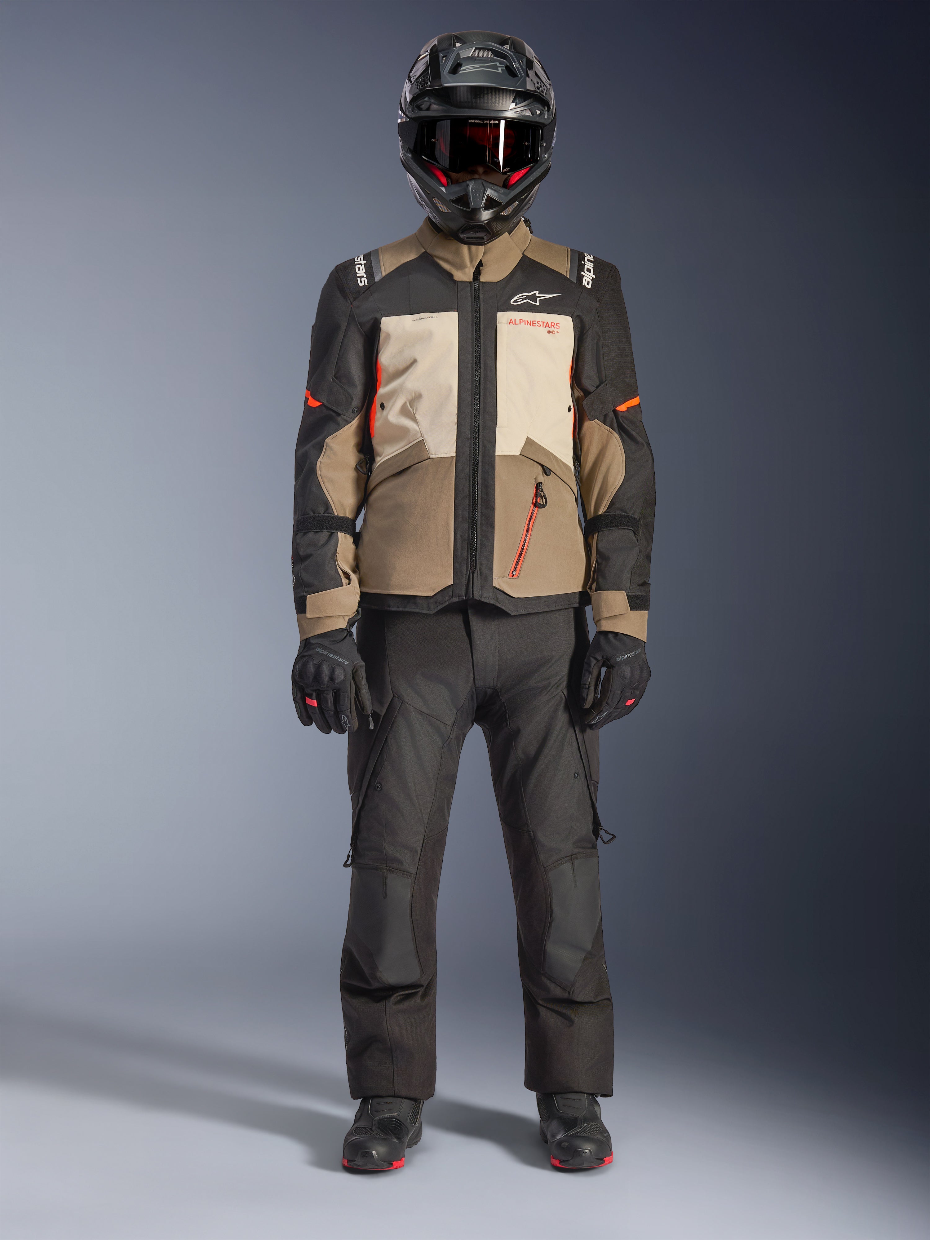 Alpinestars Andes V4 Drystar® Jacke, Walnut Mastic Black, Adventure-Tourenmotorradjacke mit hellbraunen und cremefarbenen Einsätzen und orangefarbenen Akzenten, getragen von einem Fahrer mit schwarzem Adventure-Helm, schwarzer Motorradhose und schwarzen Stiefeln, Ganzkörper-Studioansicht